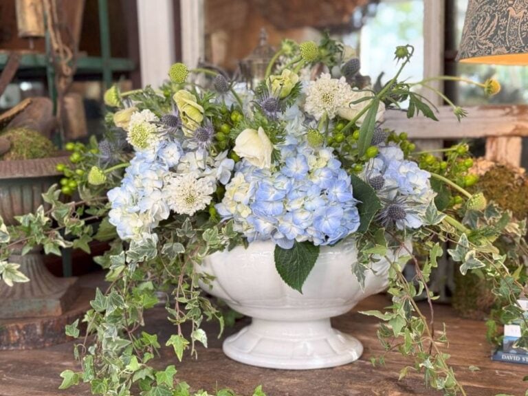 A decorative white bowl on a wooden table showcases a stunning blue hydrangea centerpiece, interspersed with white flowers, green ivy, and sheep's head blooms. The mix of greenery creates an ultimate guide to lush elegance in floral arrangements.