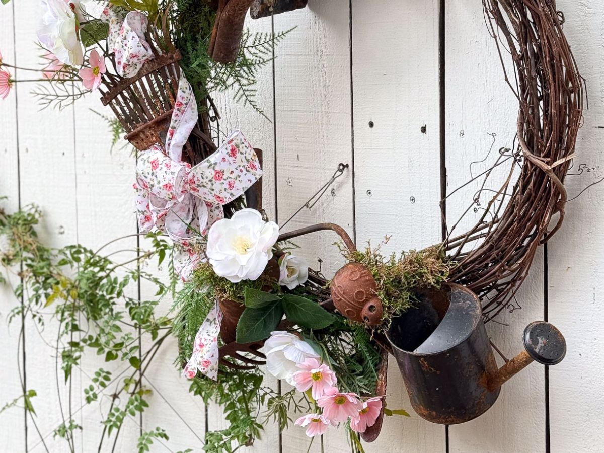 An upcycled yard tools wreath graces the white wooden wall. Rustic twigs, white and pink flowers, a floral bow, small garden tools, and a metal watering can combine for a charming garden-themed decoration, with greenery adding a fresh touch around the wreath.