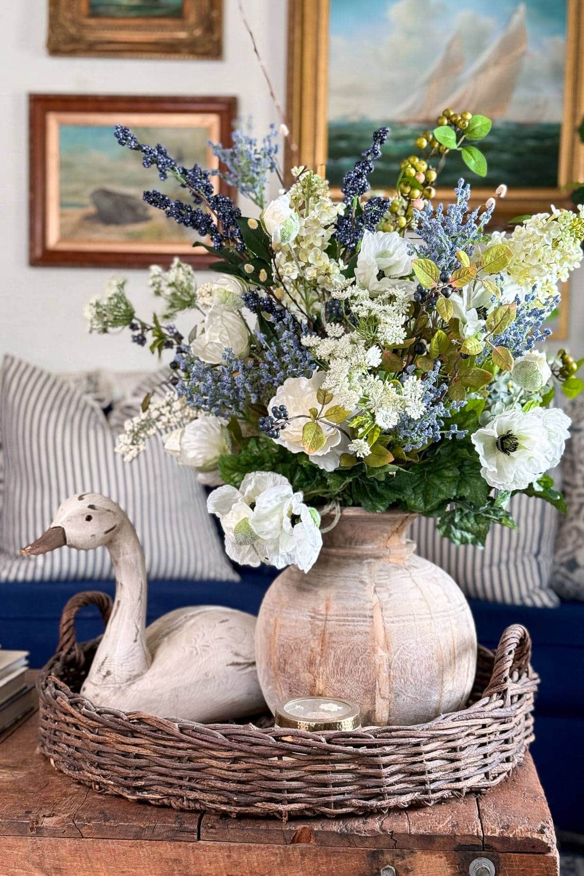A wicker tray on a wooden table holds a vase with a lush bouquet of white and blue flowers. Next to the vase is a decorative duck figure. In the background, there are striped cushions and framed paintings on the wall.