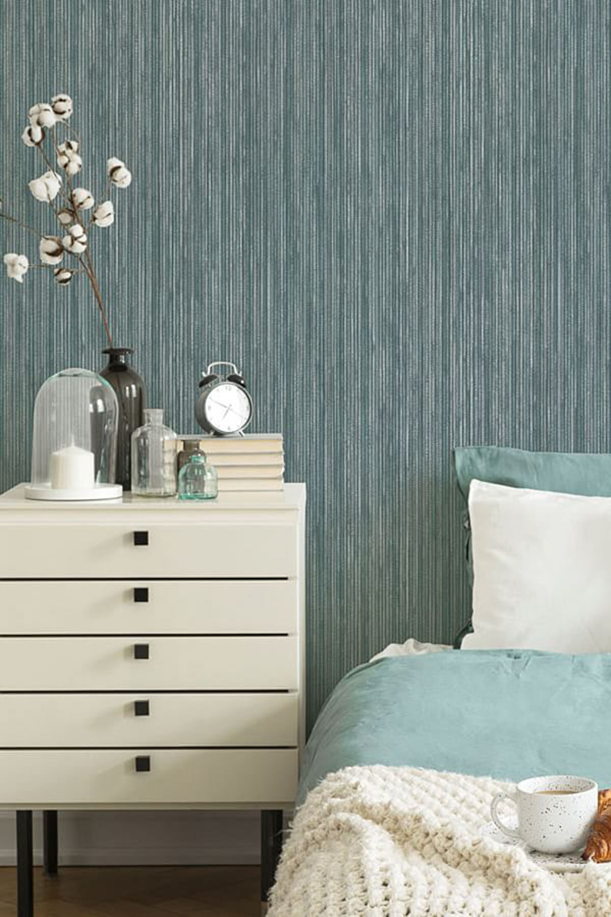 A bedroom showcases a white bedside table with books, a clock, and vases holding cotton stems. A bed dressed with a teal blanket and decorative pillows rests against textured teal wallpaper. A hot beverage sits on a knit blanket, offering a cozy touch to the updated space.