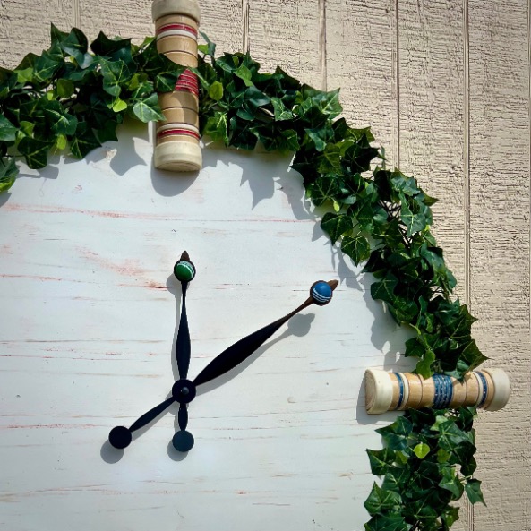 Large wall clock with a rustic look, featuring two simple black clock hands on a light wooden face. The clock is surrounded by a decorative arch made of green ivy and wooden spools, set against a vertically paneled wall.