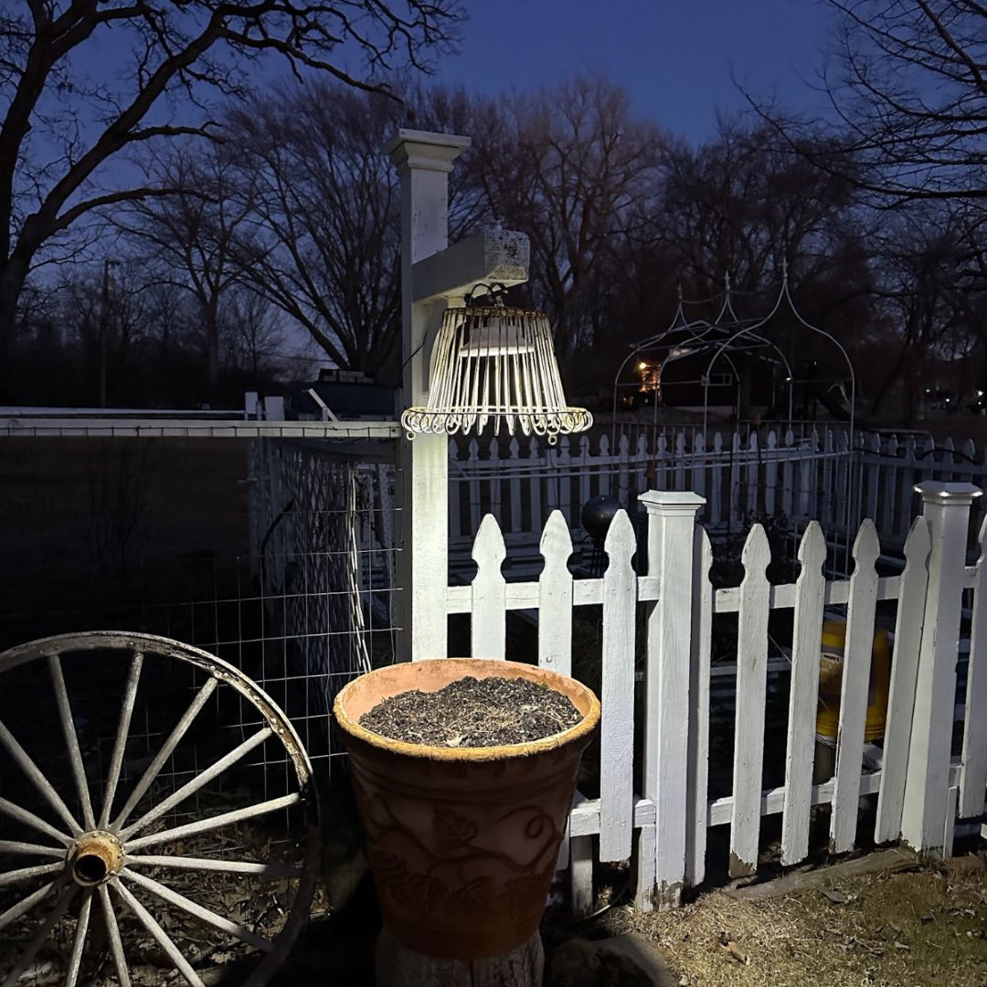 A nighttime garden scene featuring a white picket fence, a wagon wheel leaning on it, and a hanging light fixture. A large planter filled with soil is placed against the fence. Bare trees stand in the background under a dark, clear sky.