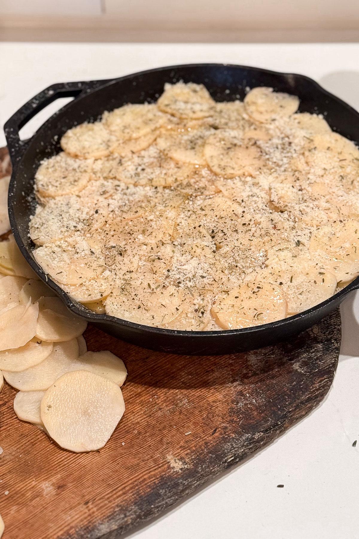 Making potato bake. A cast iron skillet filled with thinly sliced potatoes, topped with grated cheese and herbs. Some potato slices are on a wooden cutting board beside the skillet. The scene suggests preparation for baking or roasting.