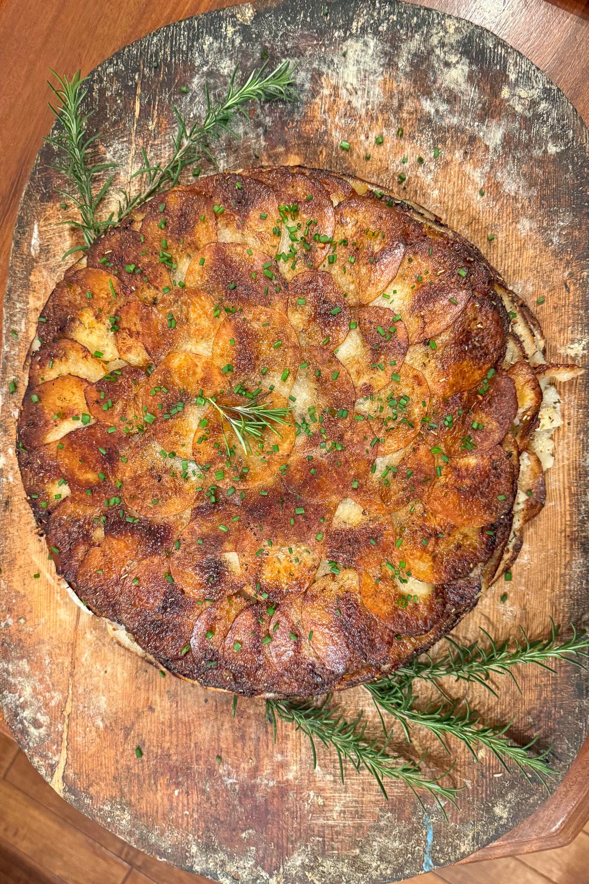 Golden-brown potato galette on a wooden serving board, garnished with fresh rosemary sprigs and chopped herbs.