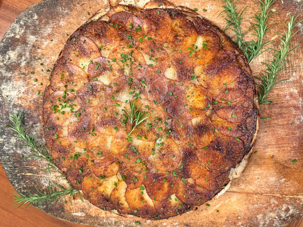 A golden-brown, crispy potato galette topped with chopped herbs on a rustic wooden board. Fresh rosemary sprigs are placed around it, enhancing its presentation.