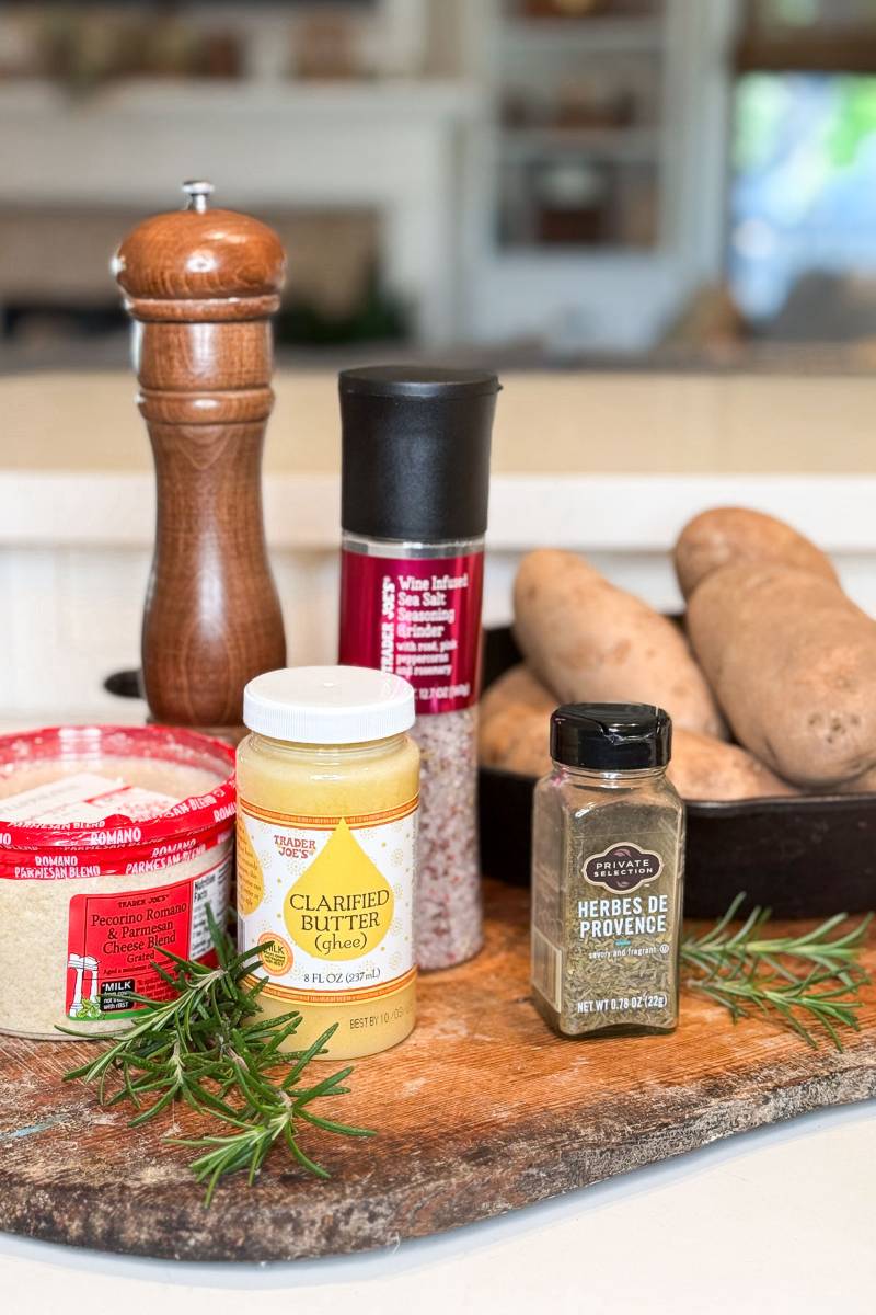 A wooden board holds a pepper grinder, Trader Joe's clarified butter, grated Pecorino Romano cheese, herbes de Provence, a salt grinder, fresh rosemary, and a black tray with sweet potatoes in a kitchen setting.