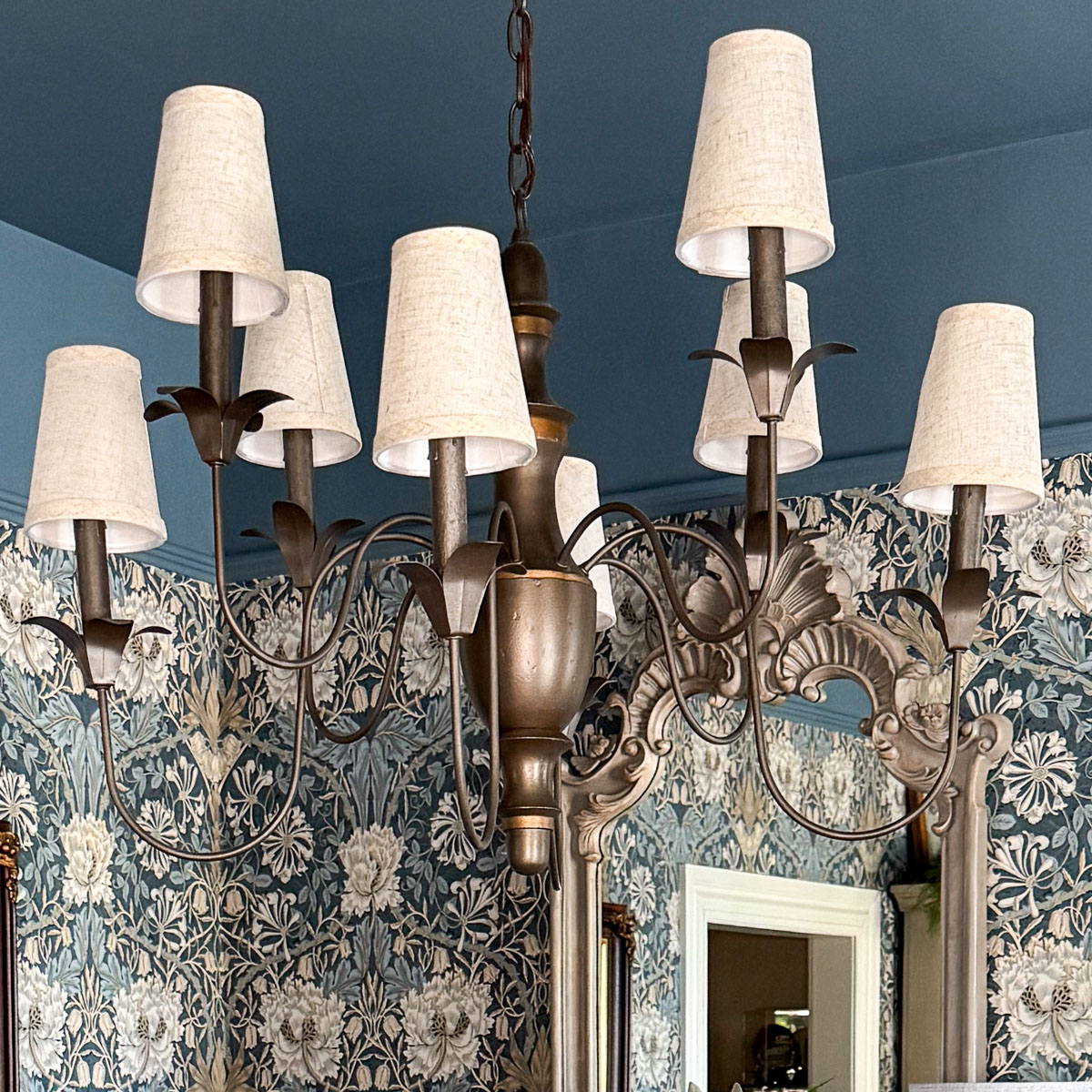 A vintage-style chandelier with eight arms and small lampshades hangs elegantly from the ceiling, adding charm to a dining room being updated with ornate floral wallpaper in blue and white. A decorative mirror peeks through, enhancing the classic ambiance.