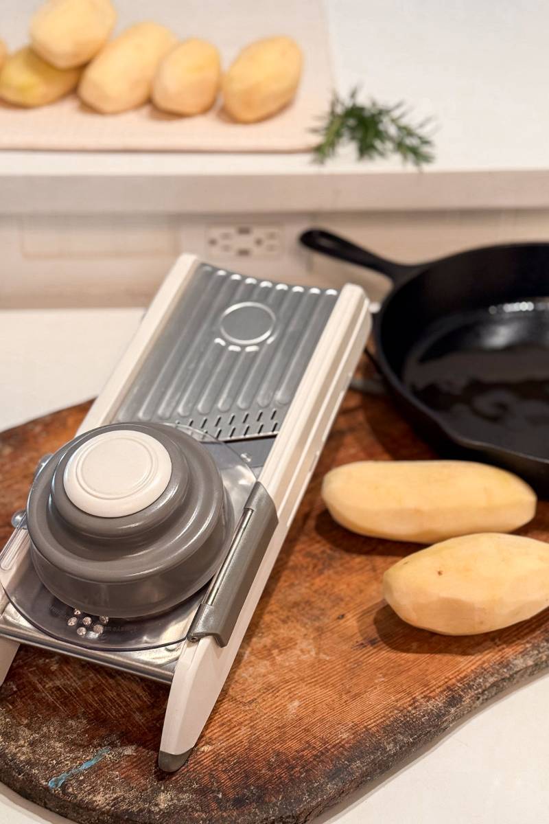 A metal mandoline slicer with a safety guard is placed on a wooden cutting board. Two peeled potatoes are beside it, with more in the background. In the corner, there's a black cast-iron skillet and a sprig of herbs.