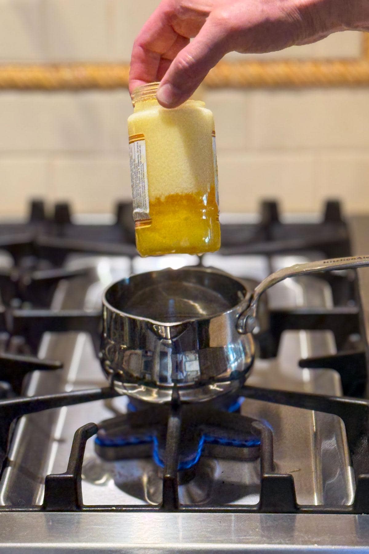 A hand holds a glass jar over a saucepan on a stove with a blue flame. The jar contains a yellow, thick substance, likely honey, with some crystallization visible inside. The stovetop is silver with black grates.