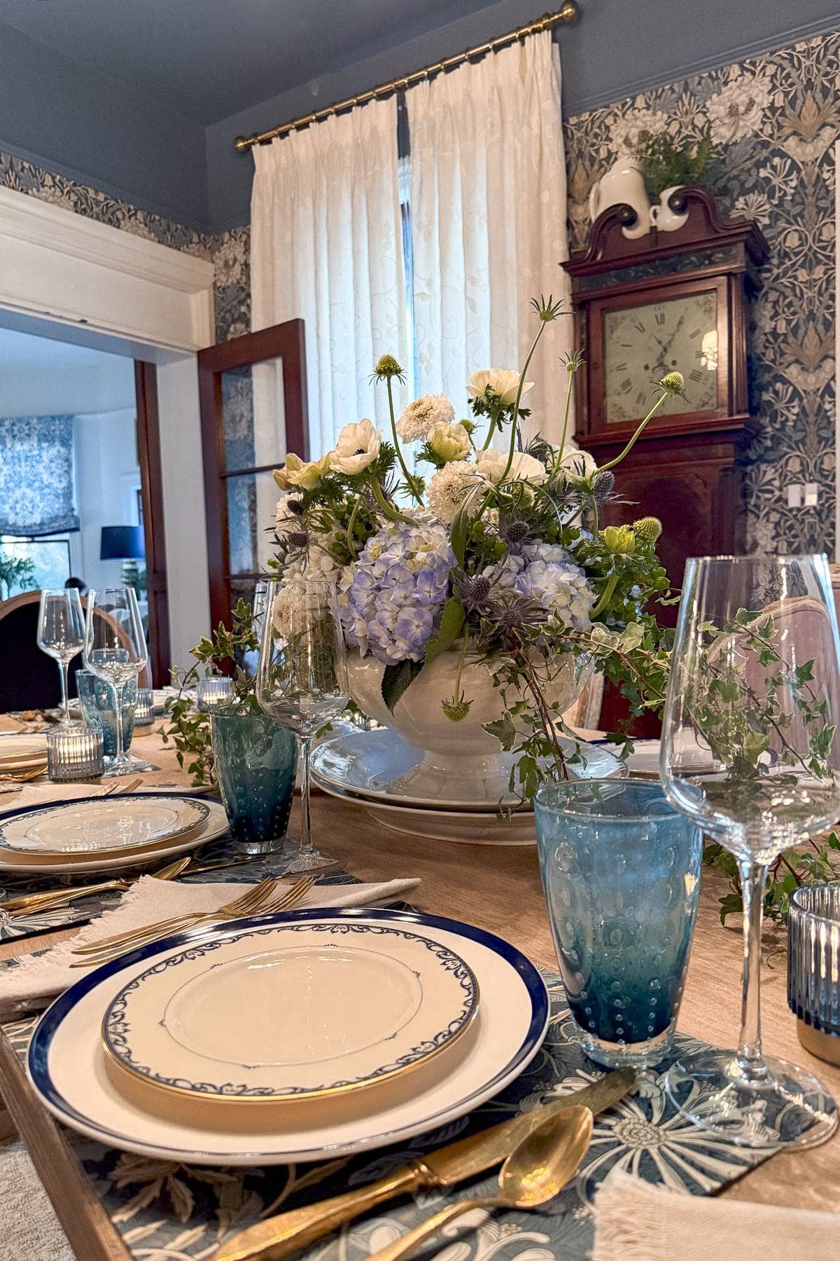 A dining table set elegantly with blue and white plates, gold cutlery, and blue glasses. A large floral centerpiece with white and blue flowers sits in the middle. The room features patterned wallpaper and a clock on the wall.