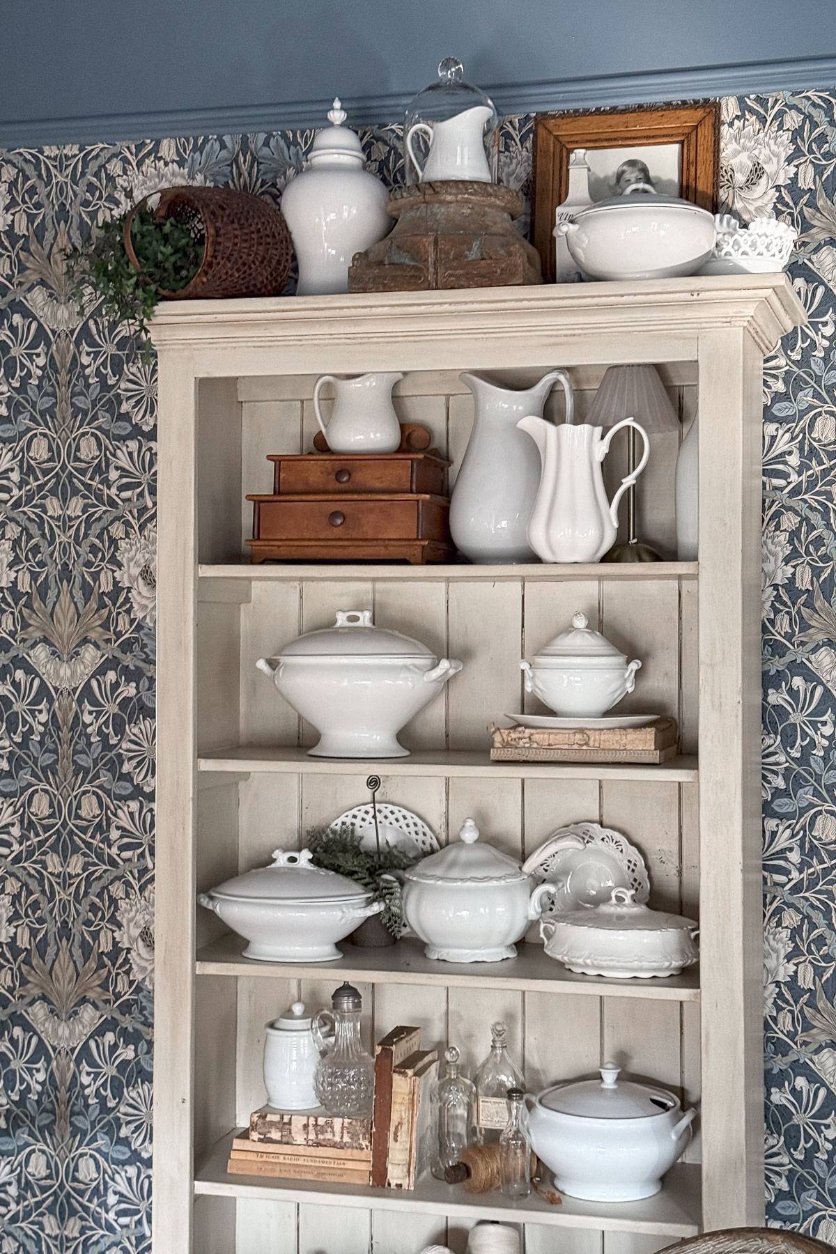 A vintage white wooden cabinet displaying an array of white ceramic dishes and pitchers. The shelves also hold small wooden boxes. The background features a floral blue and white wallpaper, adding a classic touch to the decor.