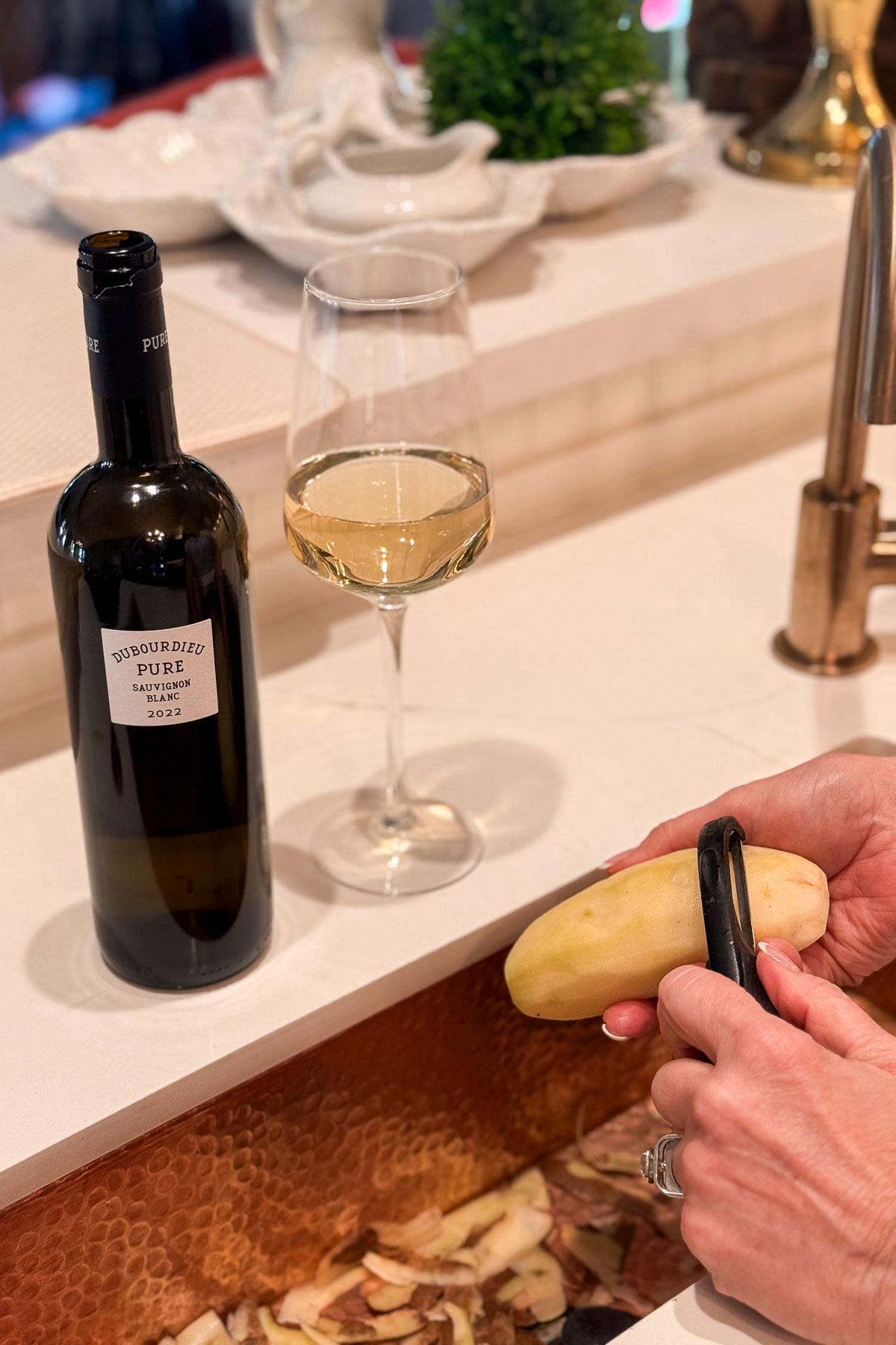 A bottle of white wine and a filled wine glass sit on a countertop. In the foreground, a person is peeling a potato over a copper sink, with peels collected beneath. A green plant and serving dishes are in the background.