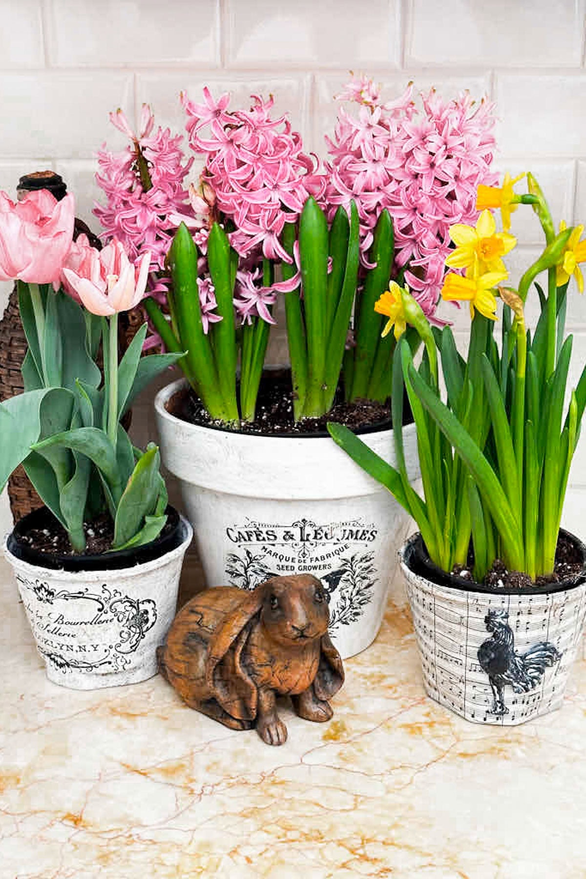 Pots of pink tulips, pink hyacinths, and yellow daffodils effortlessly decorate a marble surface. A small brown rabbit figurine sits among the plants, embodying spring decorating charm. The pots have decorative text and vintage designs.