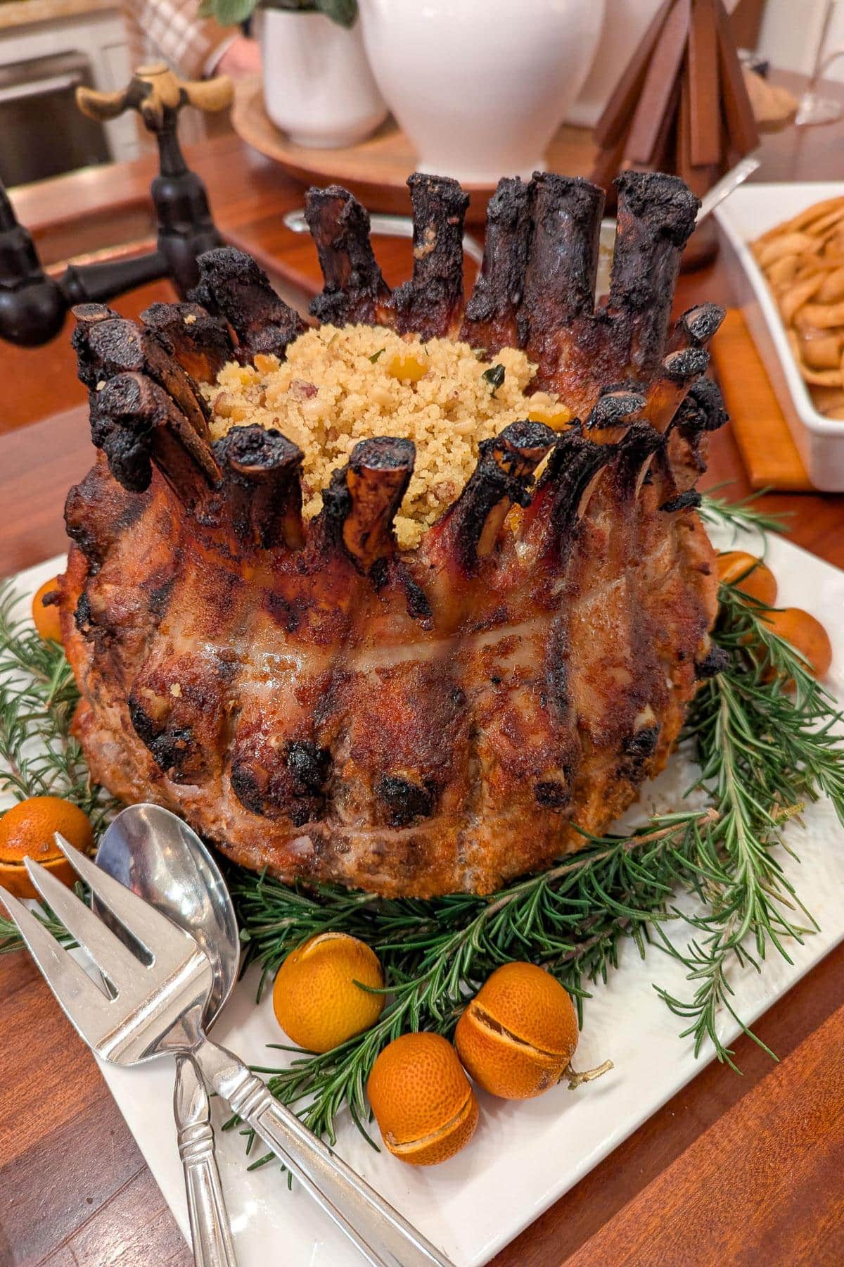 A cooked crown roast garnished with fresh rosemary and small oranges is presented on a white platter. The center of the meat is filled with a breadcrumb stuffing. Silver serving utensils are placed beside the roast.