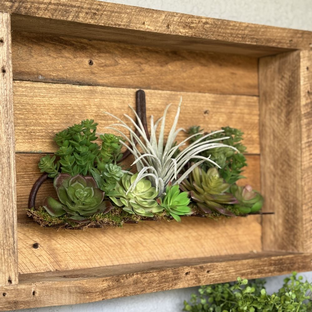 A rustic wooden frame displays a variety of green succulents and air plants, creating a vertical garden. The plants are arranged neatly, providing a contrast of textures against the wood's natural grain.