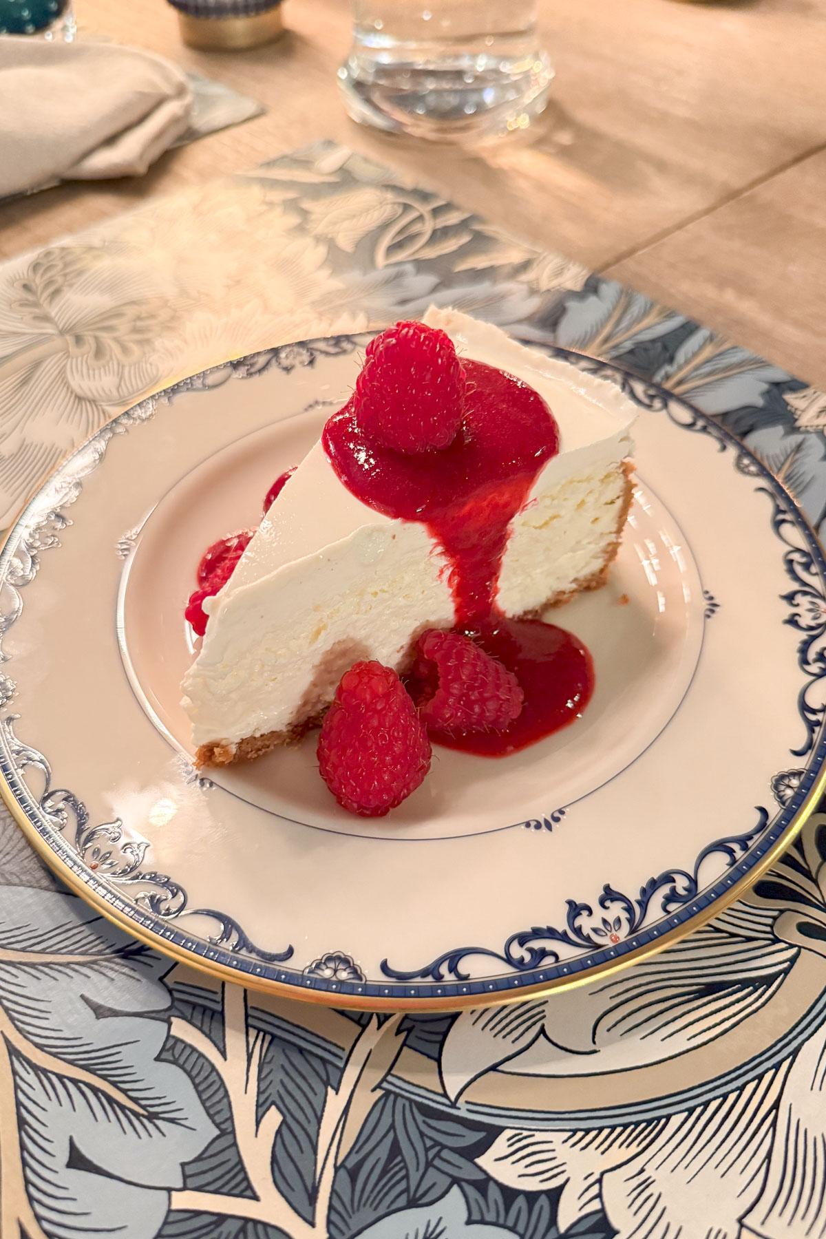 A slice of cheesecake with raspberry sauce and fresh raspberries on top, served on a decorative plate. The plate rests on a patterned table mat.