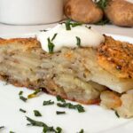 A slice of baked potato galette topped with a dollop of sour cream and sprinkled with chopped herbs on a white plate. Two whole potatoes and a sprig of rosemary are in the background.