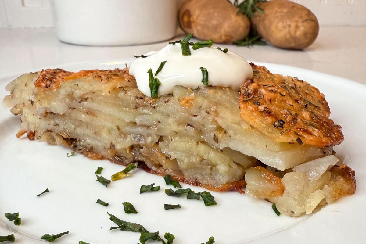 A slice of layered potato bake topped with a dollop of sour cream and sprinkled with chopped herbs on a white plate. Two whole potatoes and a sprig of rosemary are in the background.