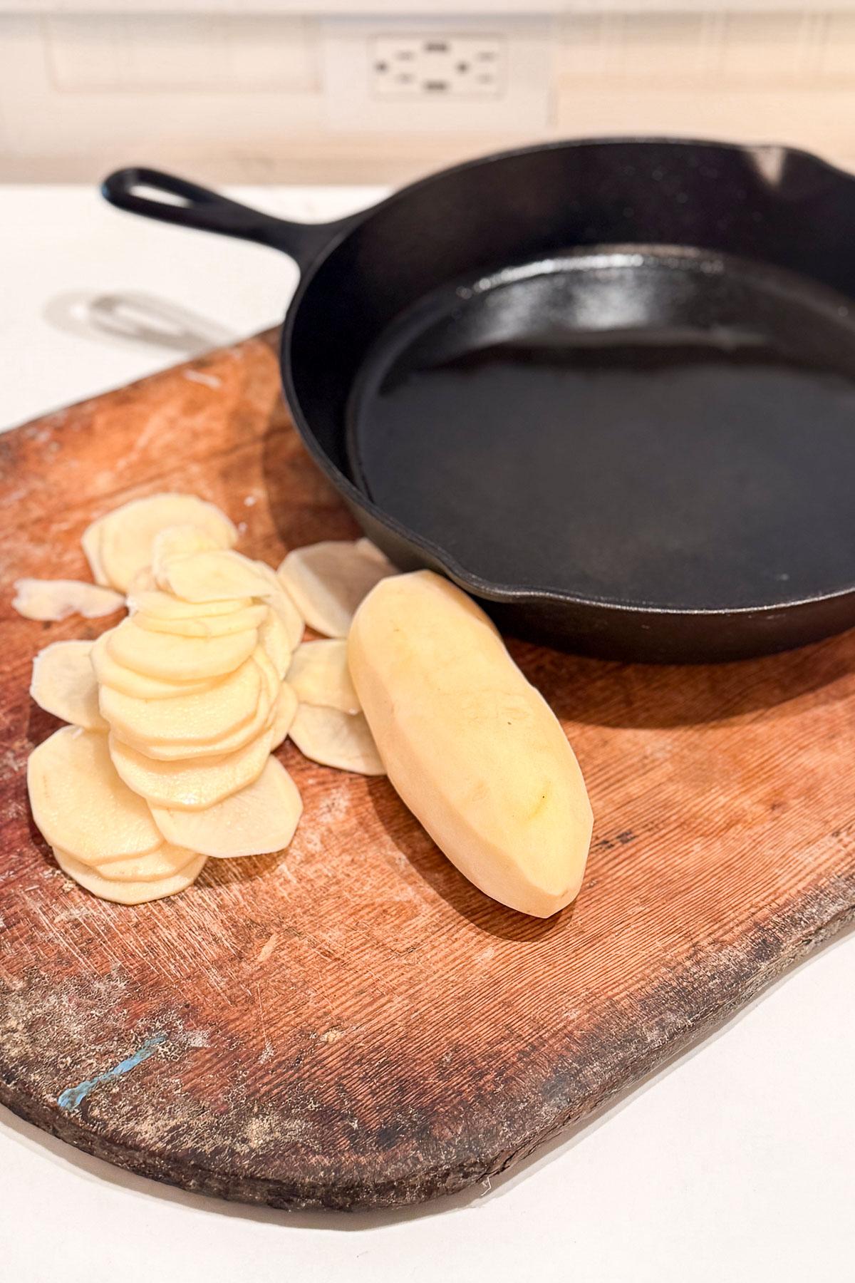 A peeled potato and several potato slices are on a worn wooden cutting board next to a black cast iron skillet. The countertop and wall socket are visible in the background.