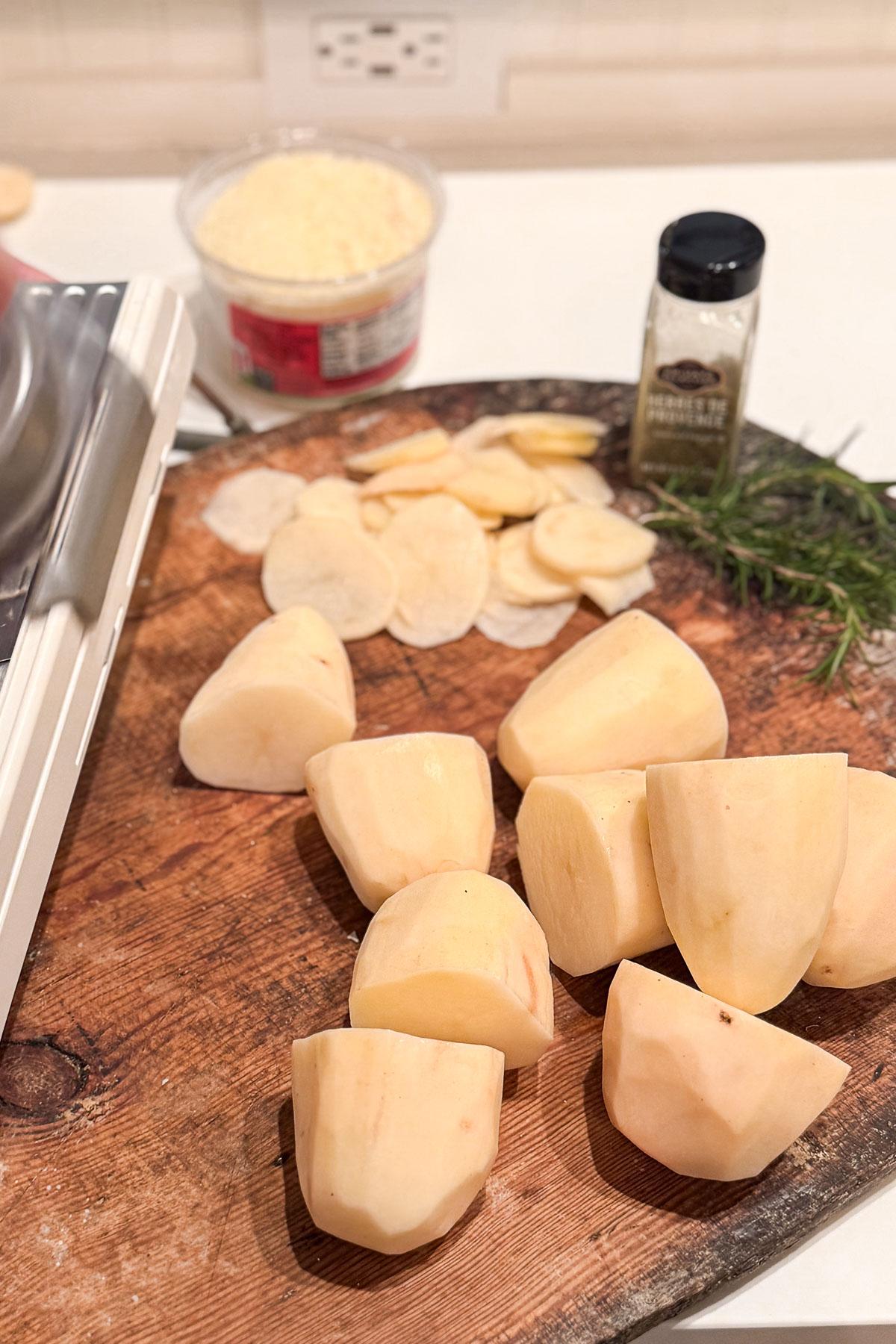 Chopped potatoes on a wooden cutting board with potato slices nearby. A tub of grated cheese, a jar of spices, and a sprig of rosemary are in the background. A mandoline slicer is partially visible on the left.