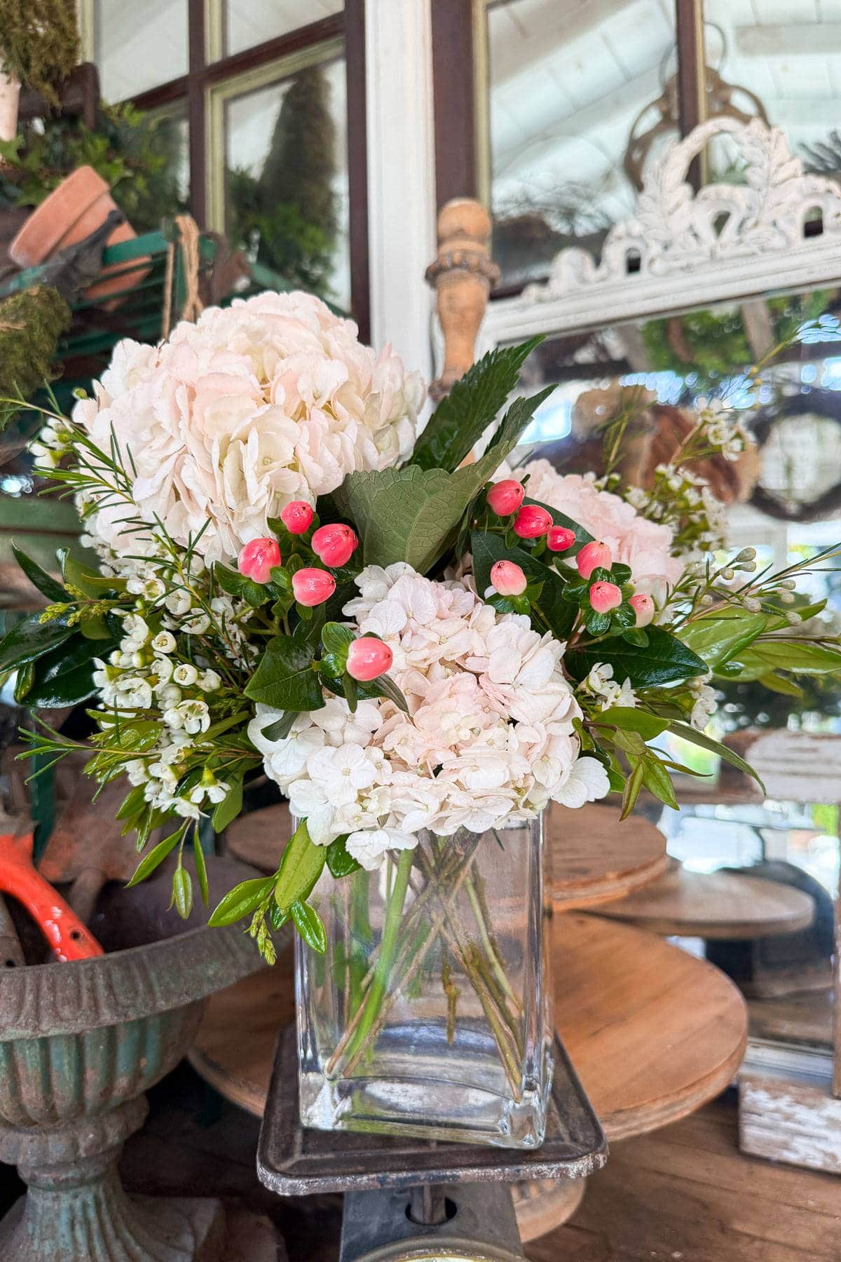 Tips for How to Keep Flower Arrangements Fresh and Beautiful for Days. A floral arrangement in a glass vase featuring white hydrangeas, pink berries, and green foliage. The vase is set on a wooden surface against a backdrop of rustic décor, including mirrors and gardening tools.
