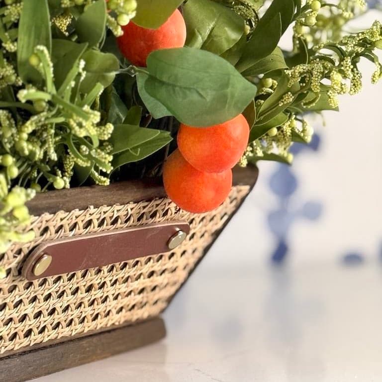 A close-up of an arrangement featuring green foliage and small, round, orange fruits in a wicker basket with a leather accent. The focus is on the vibrant greenery and fruits, with a blurred background.