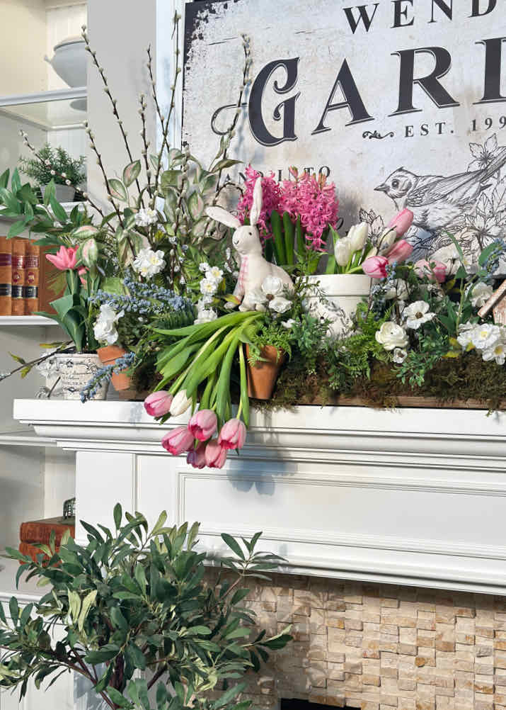 A mantel adorned for spring with fresh flowers, featuring pink tulips, white blooms, and lush greenery. A bunny figurine nests among them, while a vintage garden sign stands behind. Nearby shelves boast more plants and books for a charming spring decor.