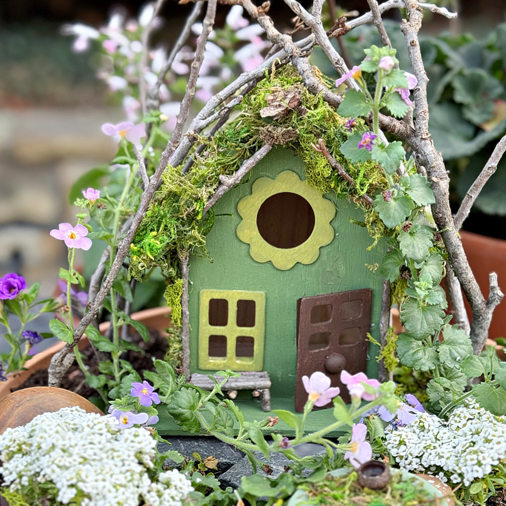 A small, whimsical green birdhouse decorated with moss and twigs, surrounded by pink and white flowers. The house features a round opening, small bench, and tiny door set in a garden scene.