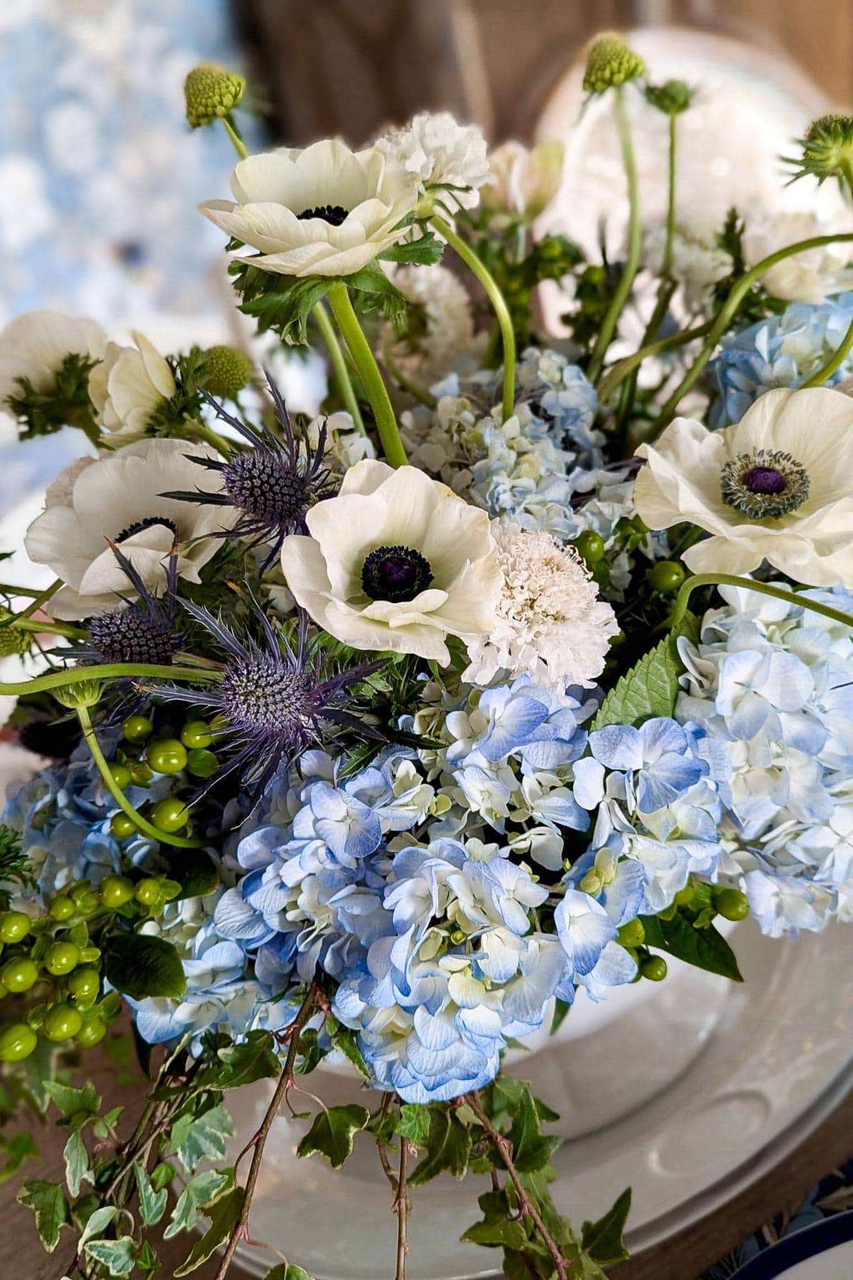 A bouquet featuring blue hydrangeas, white anemones with dark centers, purple thistles, and green berries, arranged in a white vase. The flowers are surrounded by greenery, creating a delicate and natural display.