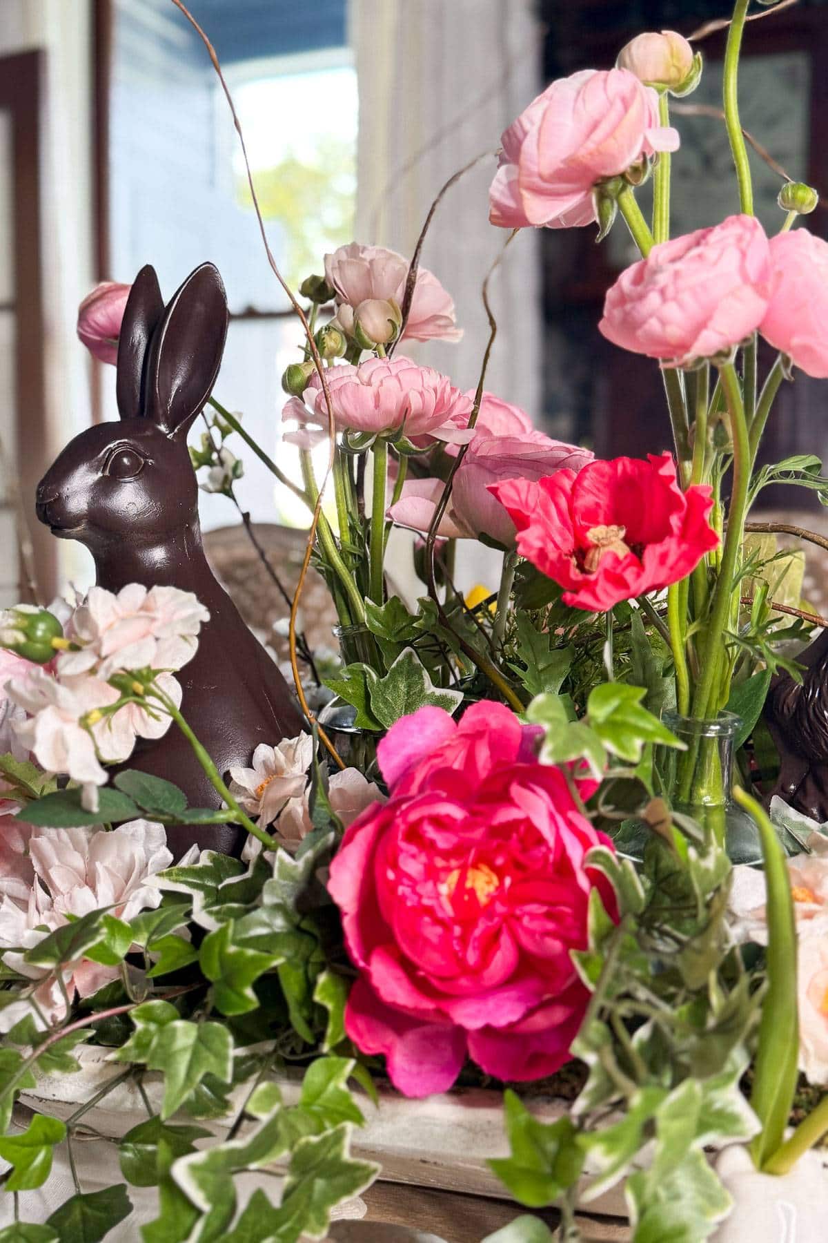 A chocolate bunny is nestled among vibrant pink roses and peonies, with green leaves and twigs adding to the arrangement. The colorful floral display is set indoors near a window.