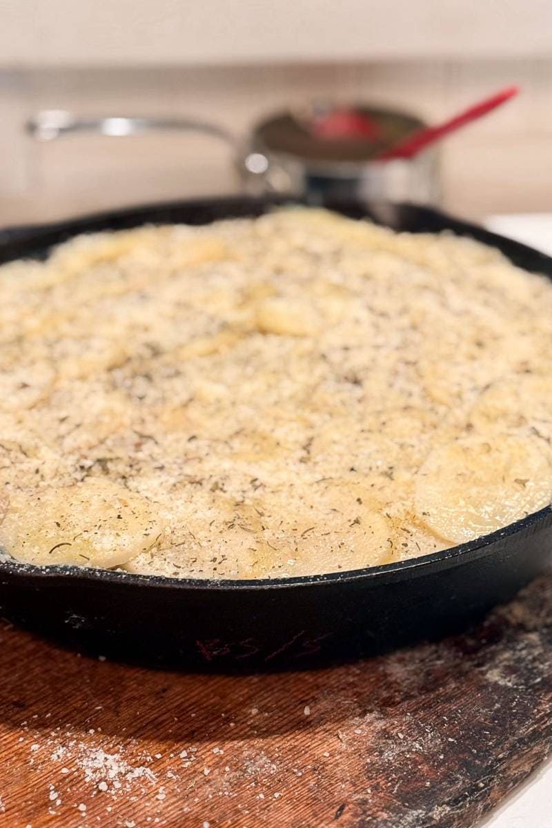 A cast iron skillet filled with scalloped potatoes topped with herbs and grated cheese, placed on a wooden cutting board. A saucepan with a red spoon is blurred in the background.