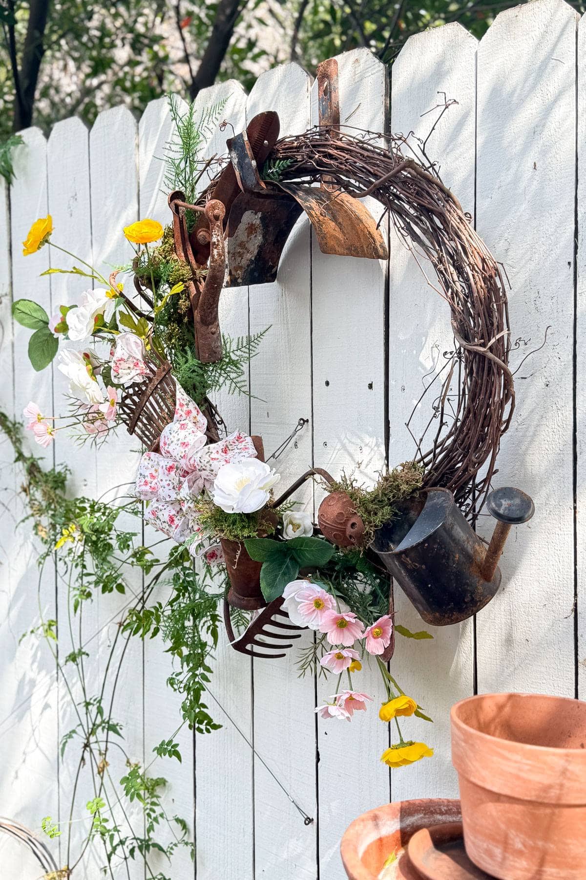 A rustic wreath made of vines, old gardening tools, and assorted flowers hangs on a white wooden fence. The wreath includes garden trowels, watering cans, and a small rake, mixed with pink and white flowers and greenery. Clay pots are nearby.