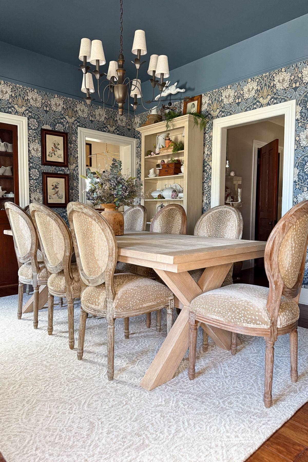 A dining room with a wooden table and eight upholstered chairs. The walls are decorated with floral wallpaper and framed art. A chandelier hangs from the ceiling. A bookshelf with decorative items stands in the background. A light rug covers the floor.