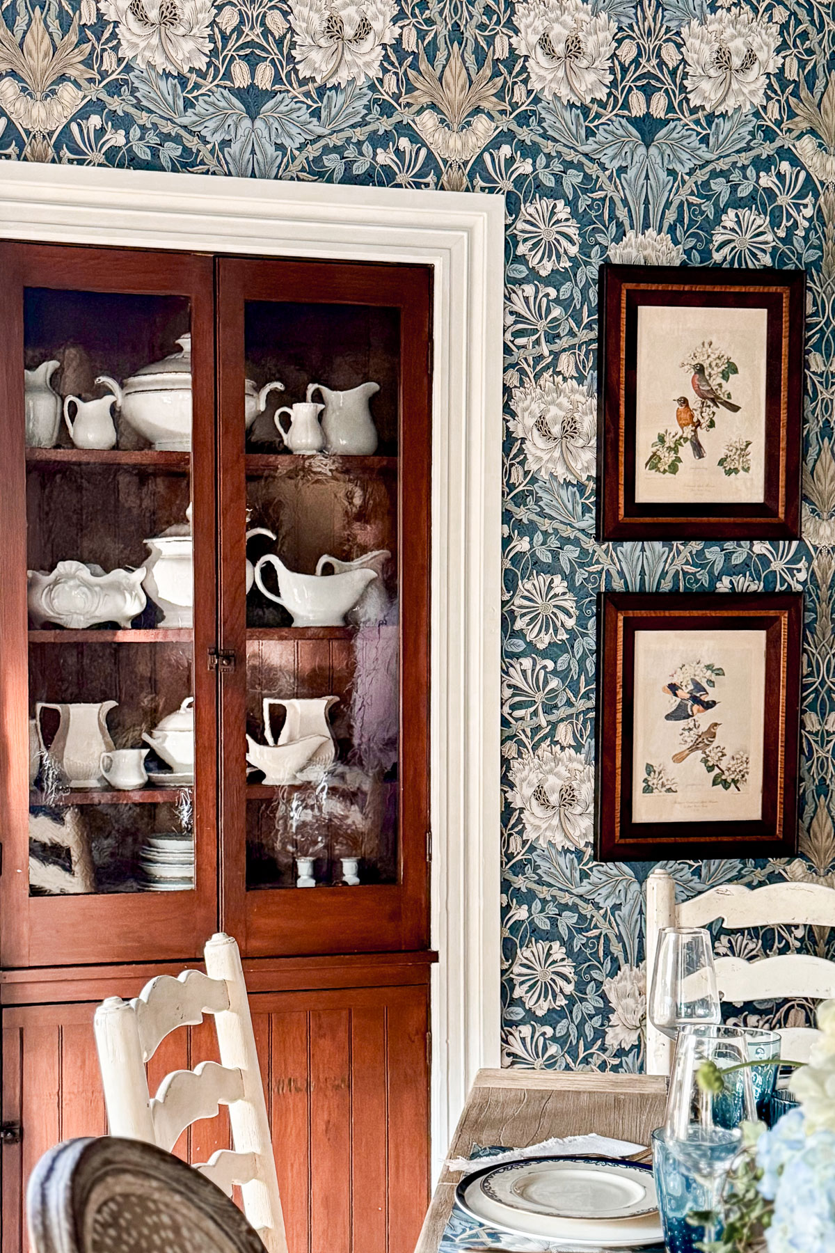 A wooden cabinet with glass doors showcases white porcelain dishes. The room, updated with floral-patterned blue wallpaper, features framed bird illustrations and a dining table elegantly set with plates and glasses.