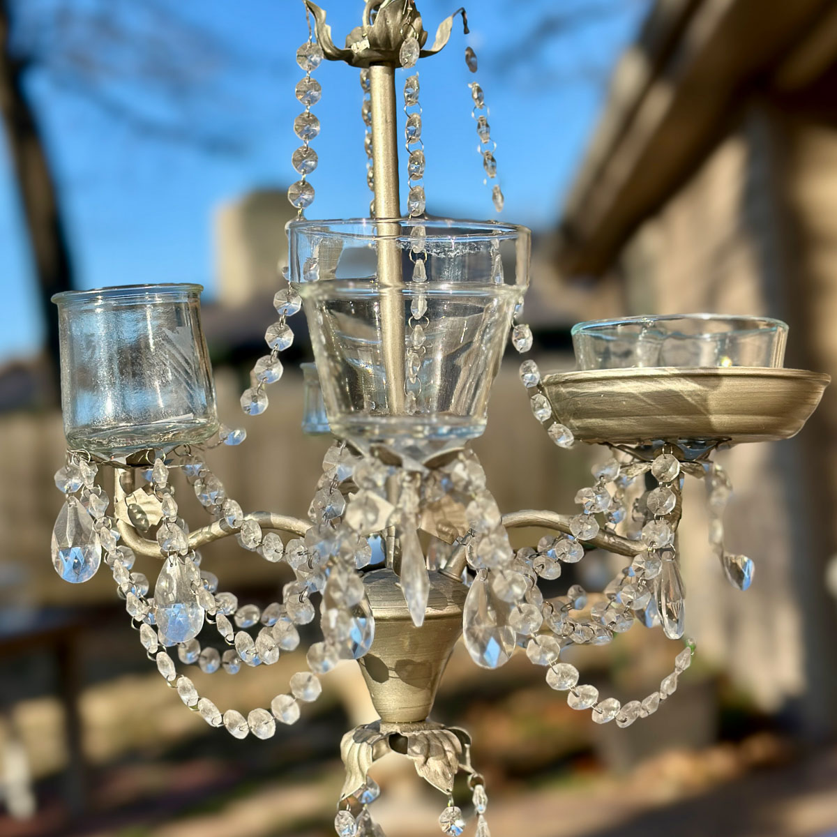 A decorative chandelier from Hand Crafted Society with dangling glass crystals and three candle holders, set against an outdoor background. The crystals glisten brightly in the light, creating a sparkling effect.