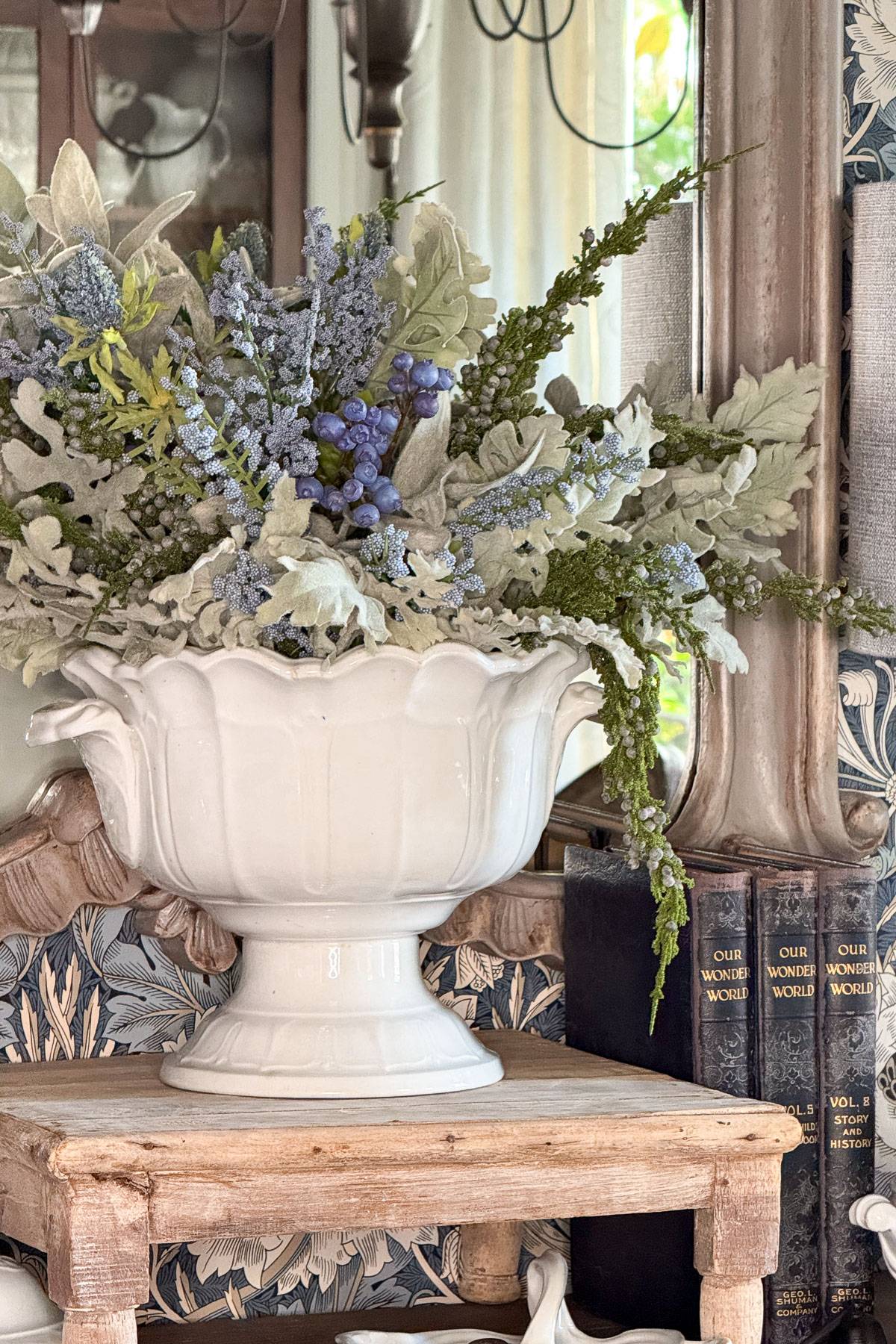 A white ceramic vase with an arrangement of green foliage and purple berries sits on a wooden stool. The background features a decorative mirror, patterned wallpaper, and a stack of books titled "Our Wonder World.