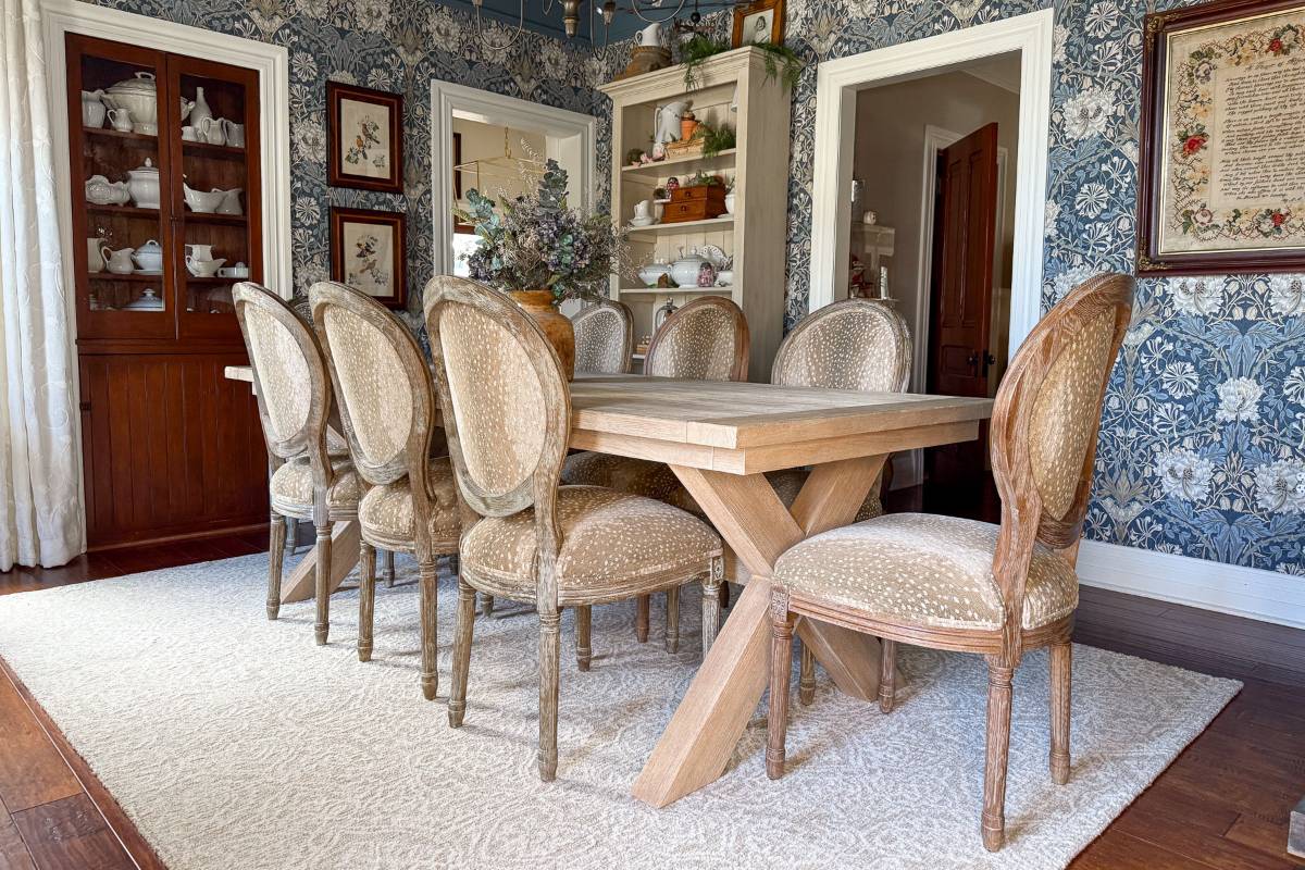 A dining room with a wooden table and eight upholstered chairs. The room features floral wallpaper and a glass cabinet filled with tea sets. A white rug lies beneath the table, and shelves with decorations are in the background.