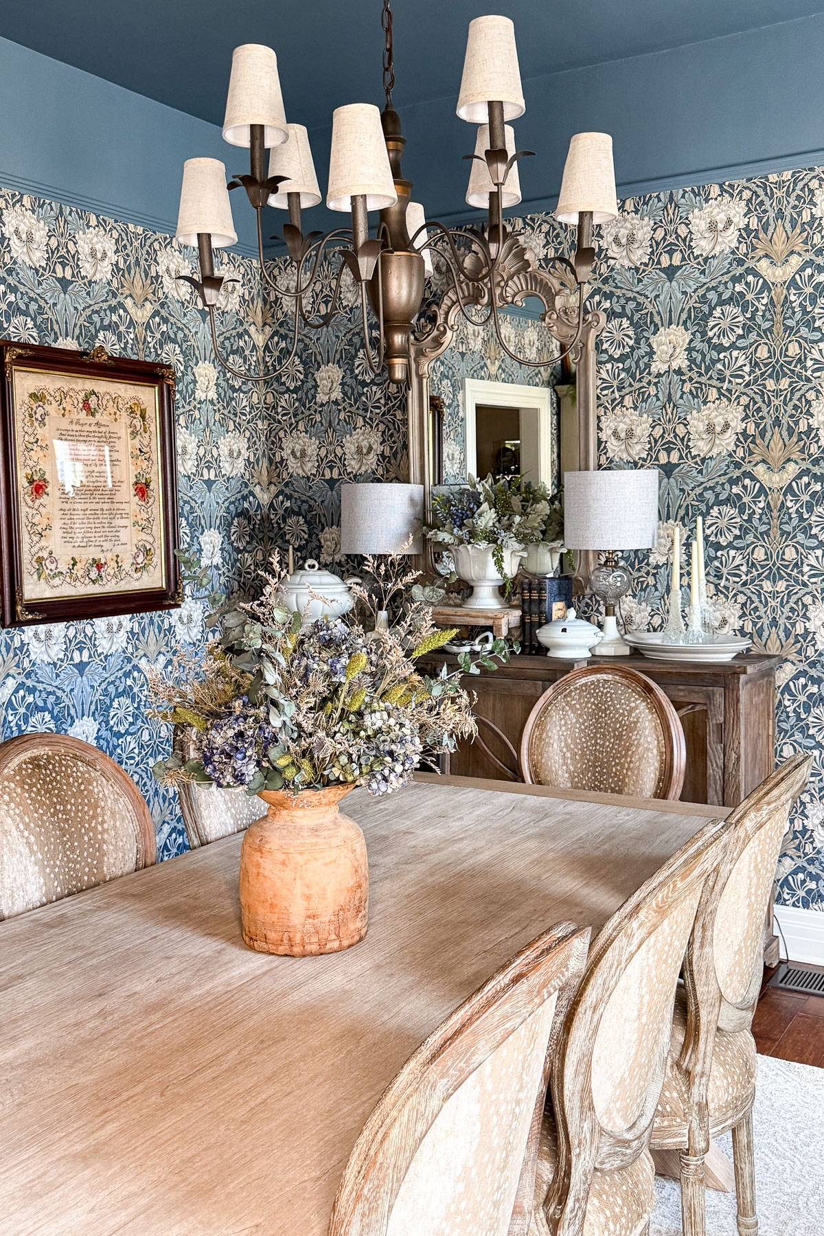 Elegant dining room with a wooden table and six upholstered chairs. A large floral arrangement in an orange vase is the centerpiece. Blue floral wallpaper decorates the room, complemented by a chandelier and a sideboard with lamps and decor items.