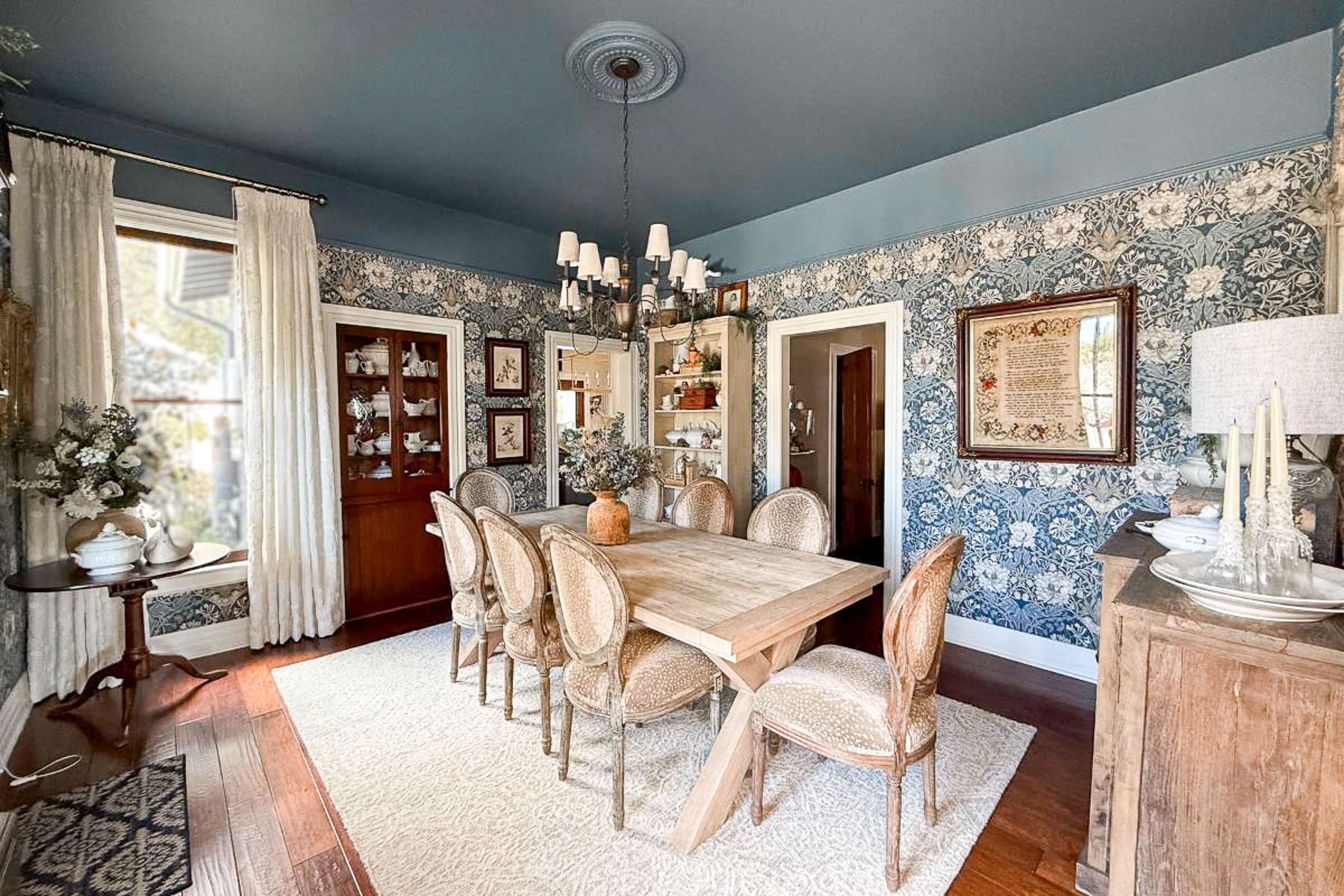 A cozy dining room with a wooden table and six wicker chairs on a light rug. The walls, updated with blue floral wallpaper, are adorned with framed art. A dark chandelier hangs from the blue ceiling, while a window with white curtains brings in light.