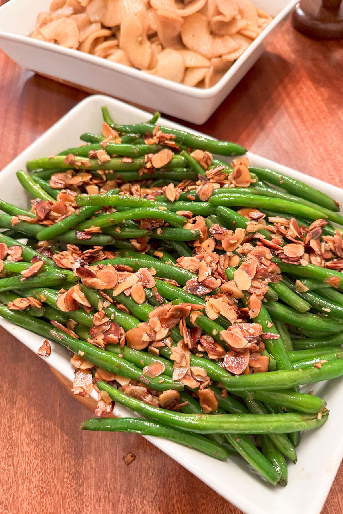 A white rectangular dish filled with green beans topped with sliced, toasted almonds. In the background, a square bowl contains thinly sliced, round pieces of a light-colored vegetable on a wooden table.