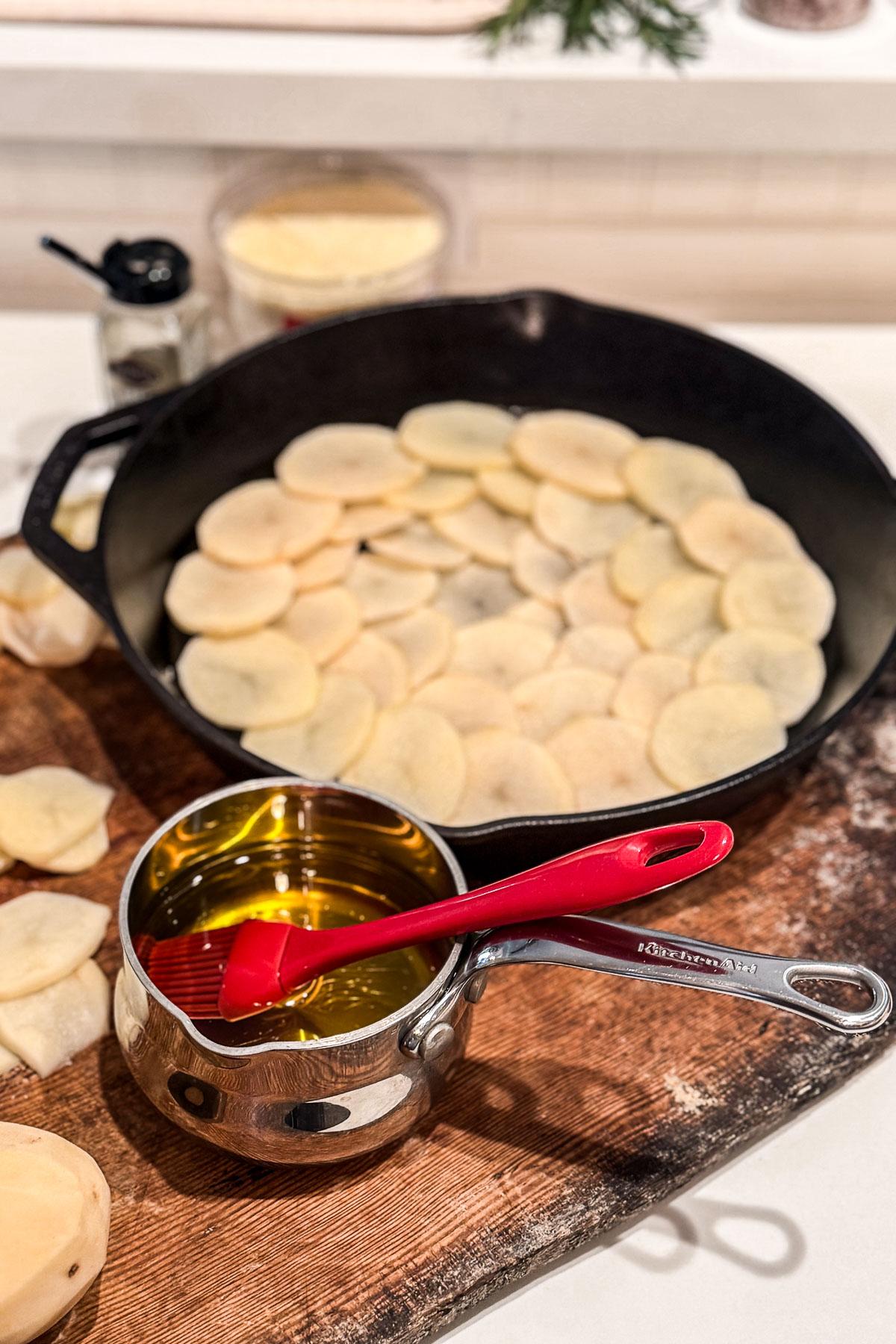 Sliced potatoes are arranged in a large black skillet on a wooden surface. A small pot filled with oil and a red silicone brush are beside it. Ingredient containers are in the background.