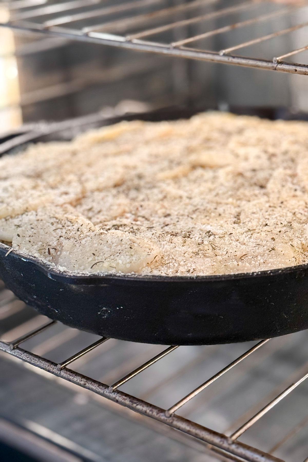 A cast iron skillet filled with a crumb-topped dish is baking on the middle rack of an oven. The dish appears golden and crispy with a textured surface, suggesting a savory topping of herbs and breadcrumbs.