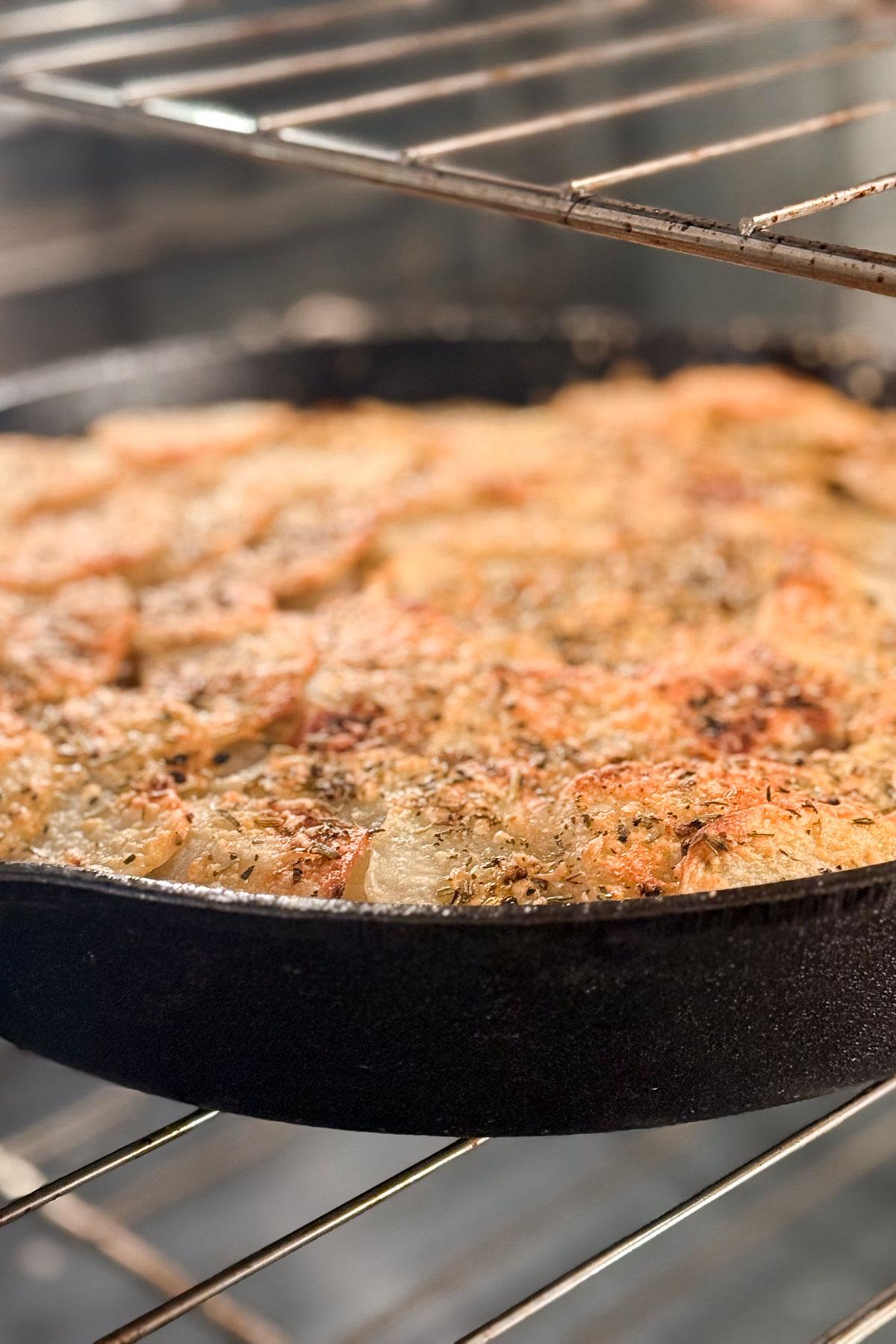 A close-up of a skillet filled with layered potato bake topped with herbs, sitting on an oven rack. The vegetables appear golden brown and crispy, suggesting they are well-cooked. The oven's interior is visible in the background.