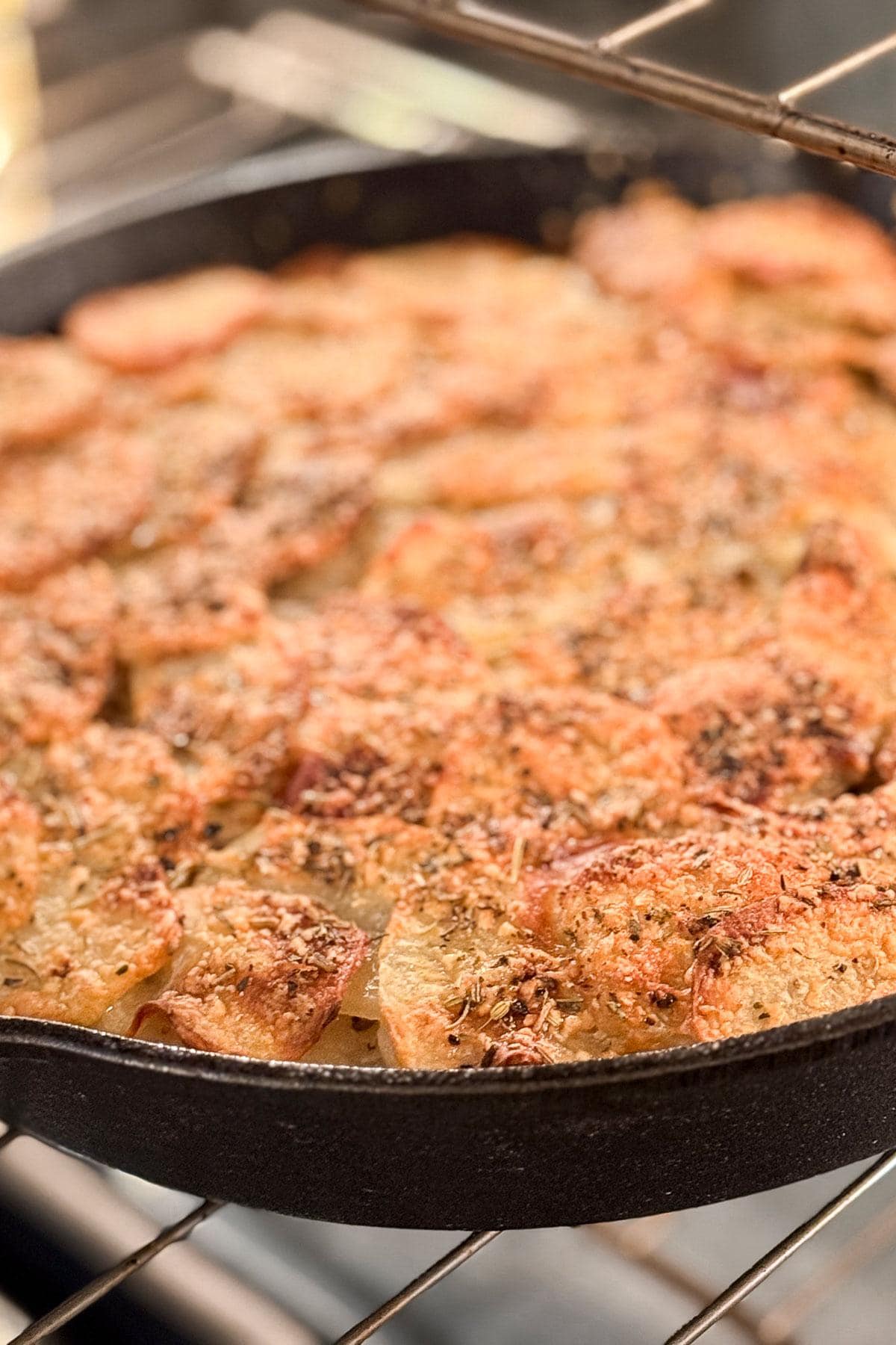 A skillet filled with golden-brown potatoes roasting in an oven. The potatoes are sliced and topped with herbs, forming a crispy layer on top. The skillet is positioned on an oven rack.