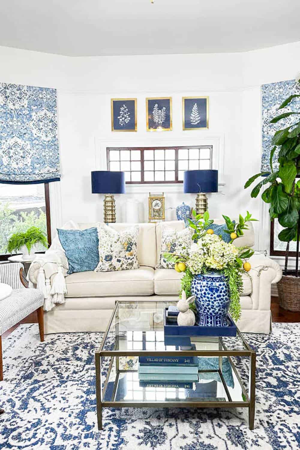 A cozy living room features a white sofa with blue-patterned cushions, a glass coffee table with books and plants, and blue and white floral curtains. Artwork adorns the walls like updating a dining room with wallpaper. A large plant graces the corner, complemented by blue table lamps on the console.