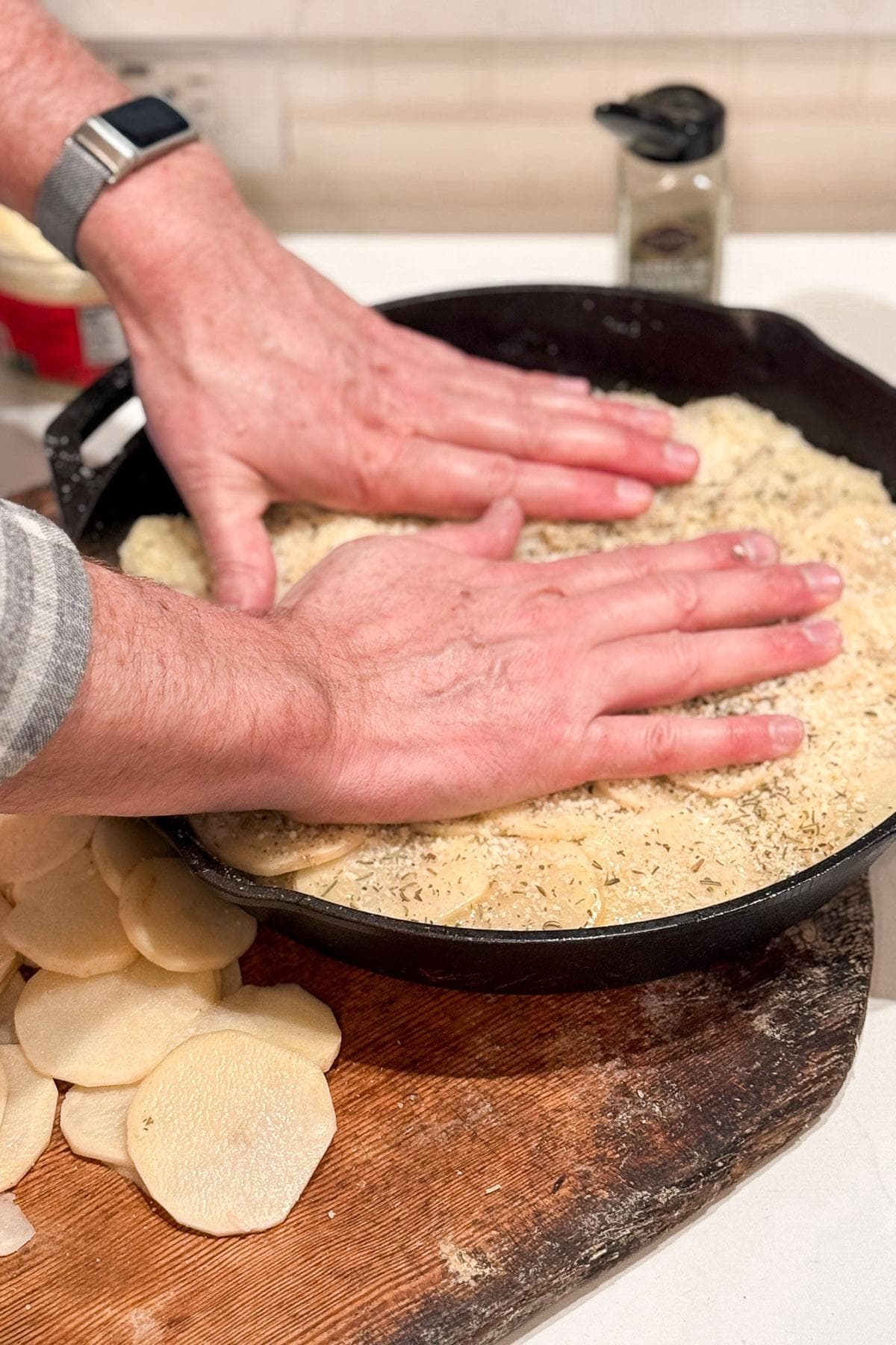 Making layered potato bake. Close-up of hands pressing a mixture of sliced potatoes and seasonings into a black cast iron skillet. A wooden board with more potato slices and a spice container are nearby. The person is wearing a watch on their left wrist.