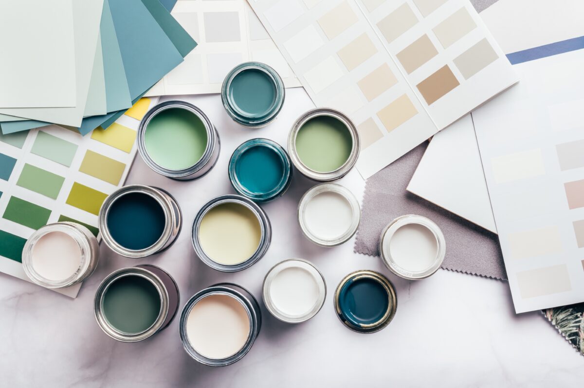 Open paint cans in shades of blue, green, and neutral tones are spread out on a surface. Color swatches and fabric samples surround them, suggesting an updating project. Could it be new wallpaper or a refreshed dining room design?.