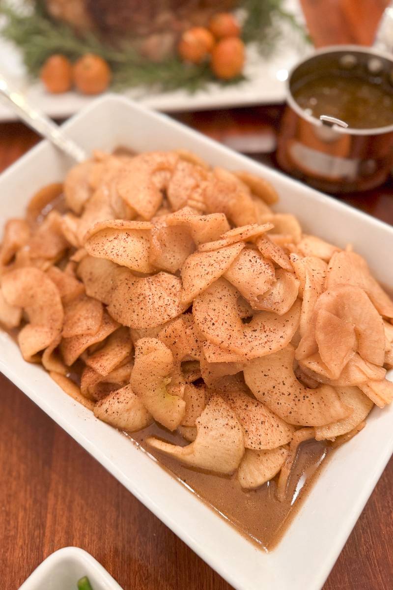 A rectangular white dish filled with caramelized apple slices sprinkled with cinnamon, served in a brown sauce. The dish is on a wooden table with a sauce container in the background.