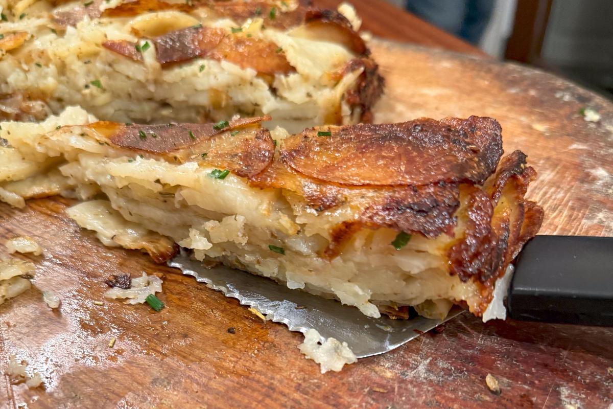 A slice of crispy, golden-brown layered potato gratin bake on a wooden cutting board. The layers of thinly sliced potatoes are visible, with a sprinkle of herbs. A knife partially slides under the slice, with bits of potato scattered around.