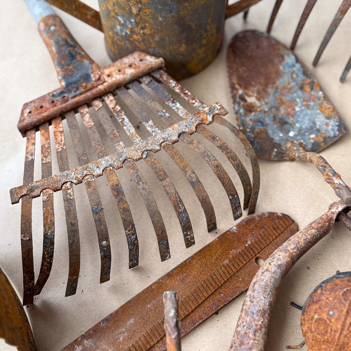 Close-up of rusty gardening tools, including a rake, spade, and watering can, arranged on a beige surface. These treasures from the Hand Crafted Society display heavy corrosion, showcasing their age and wear with pride.